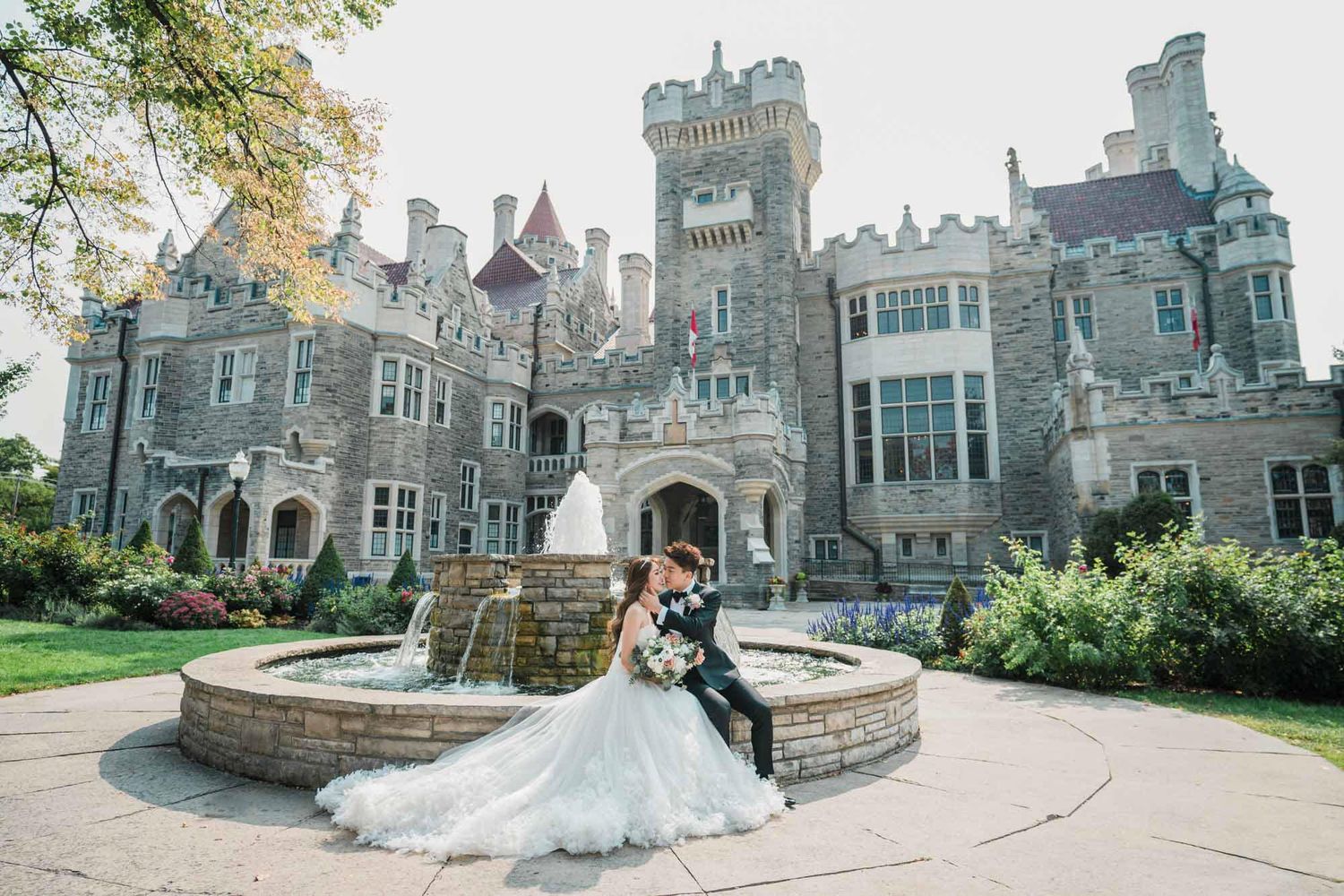 Wedding couple shares a romantic kiss by a stone fountain in front of a majestic castle venue.