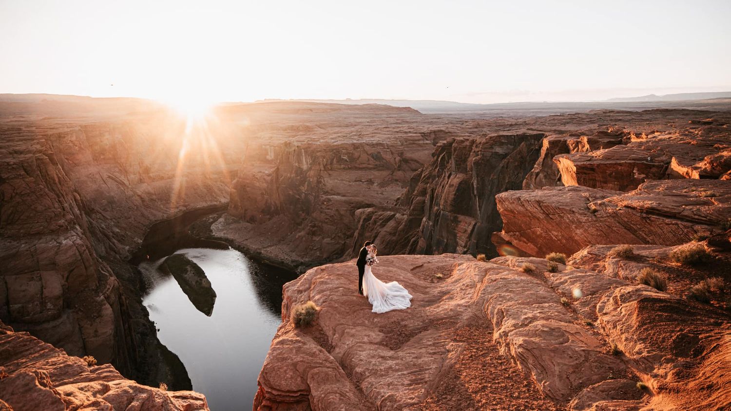 Bride in flowing white dress standing on Horseshoe Bend cliff edge at stunning golden sunset.