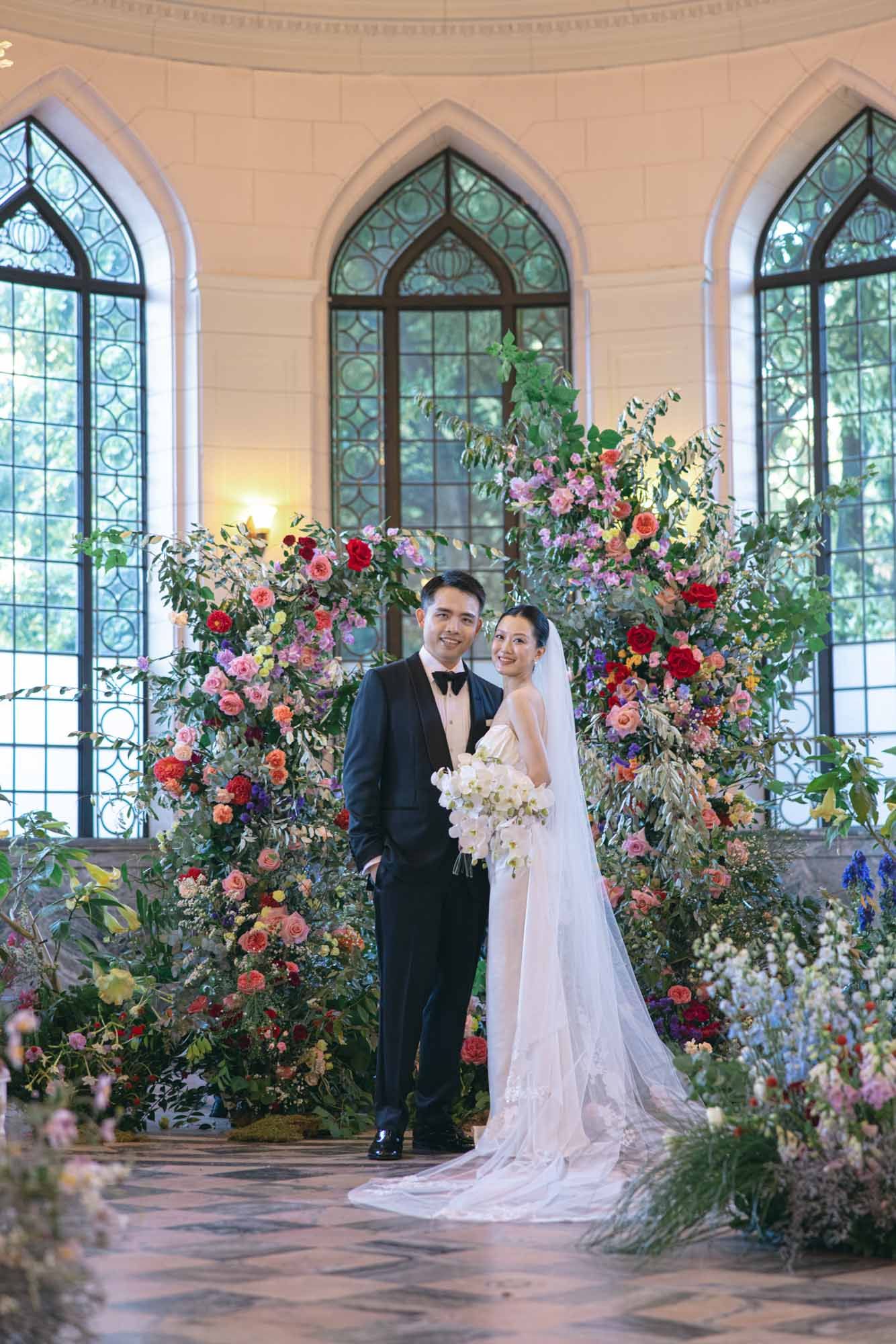 Couple poses before stunning colorful floral arch in Gothic chapel wedding ceremony venue.