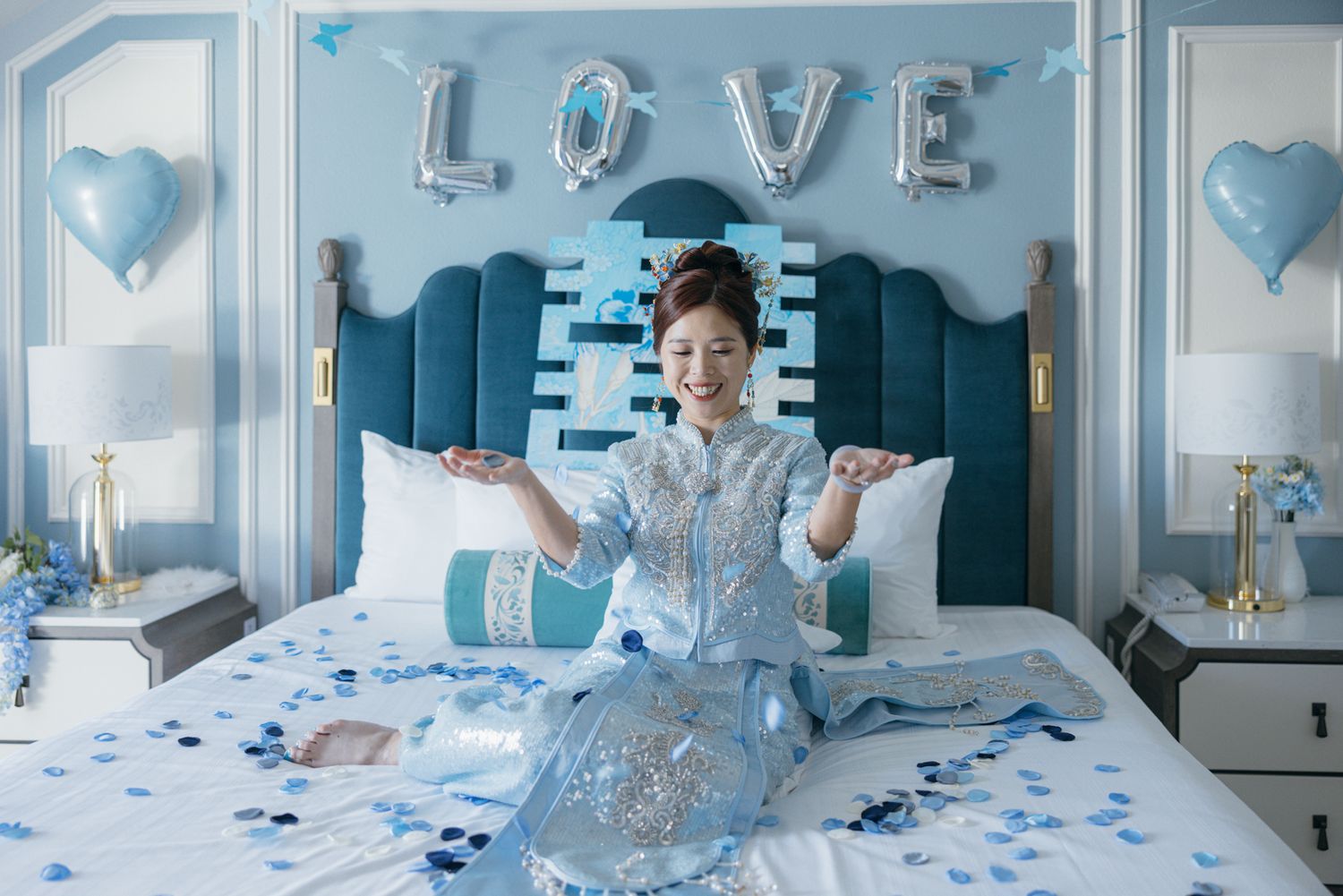 Bride in blue qipao sits on decorated bed surrounded by confetti under LOVE balloon letters.