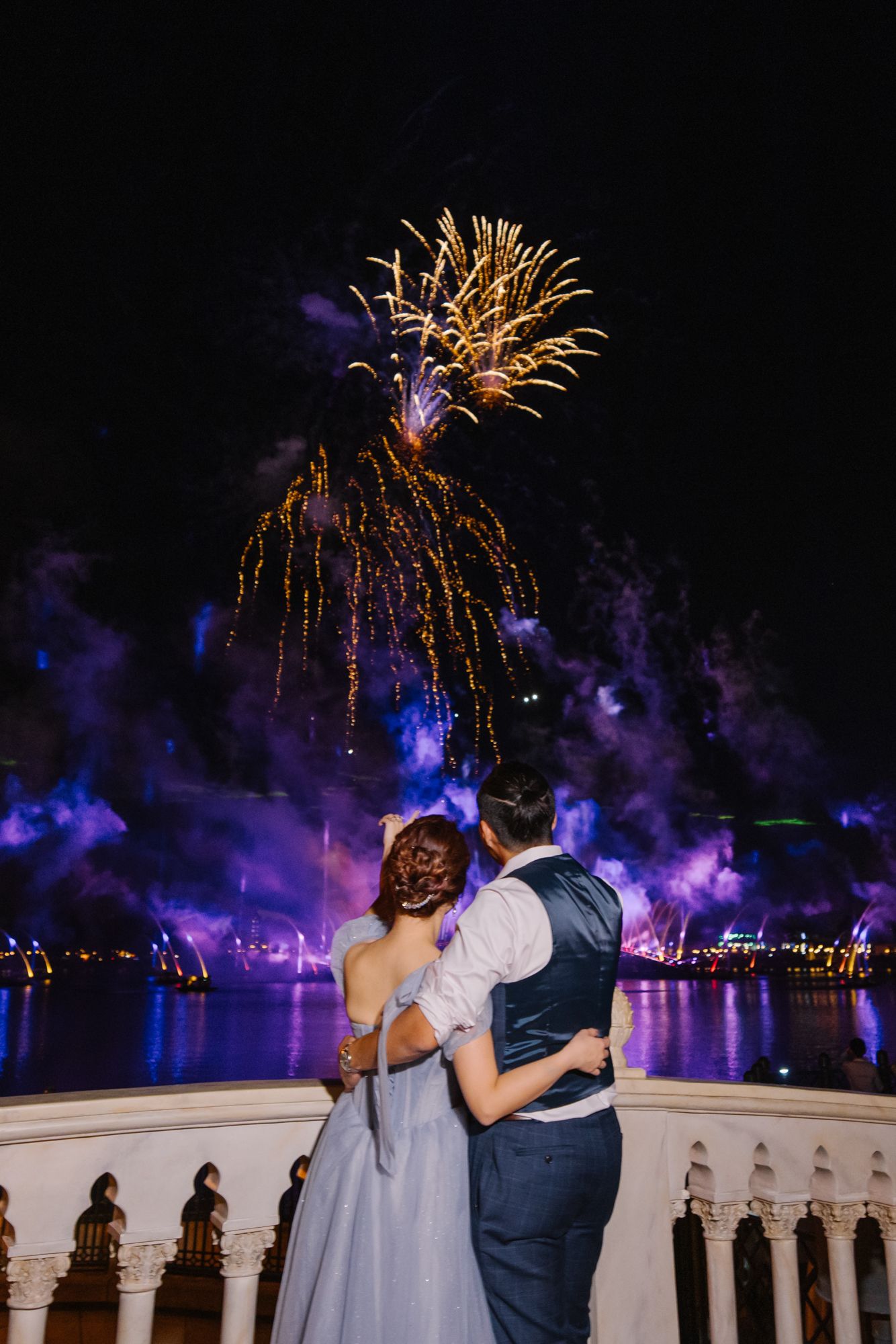 Newlyweds embrace while watching colorful fireworks display over water at Disney wedding reception.