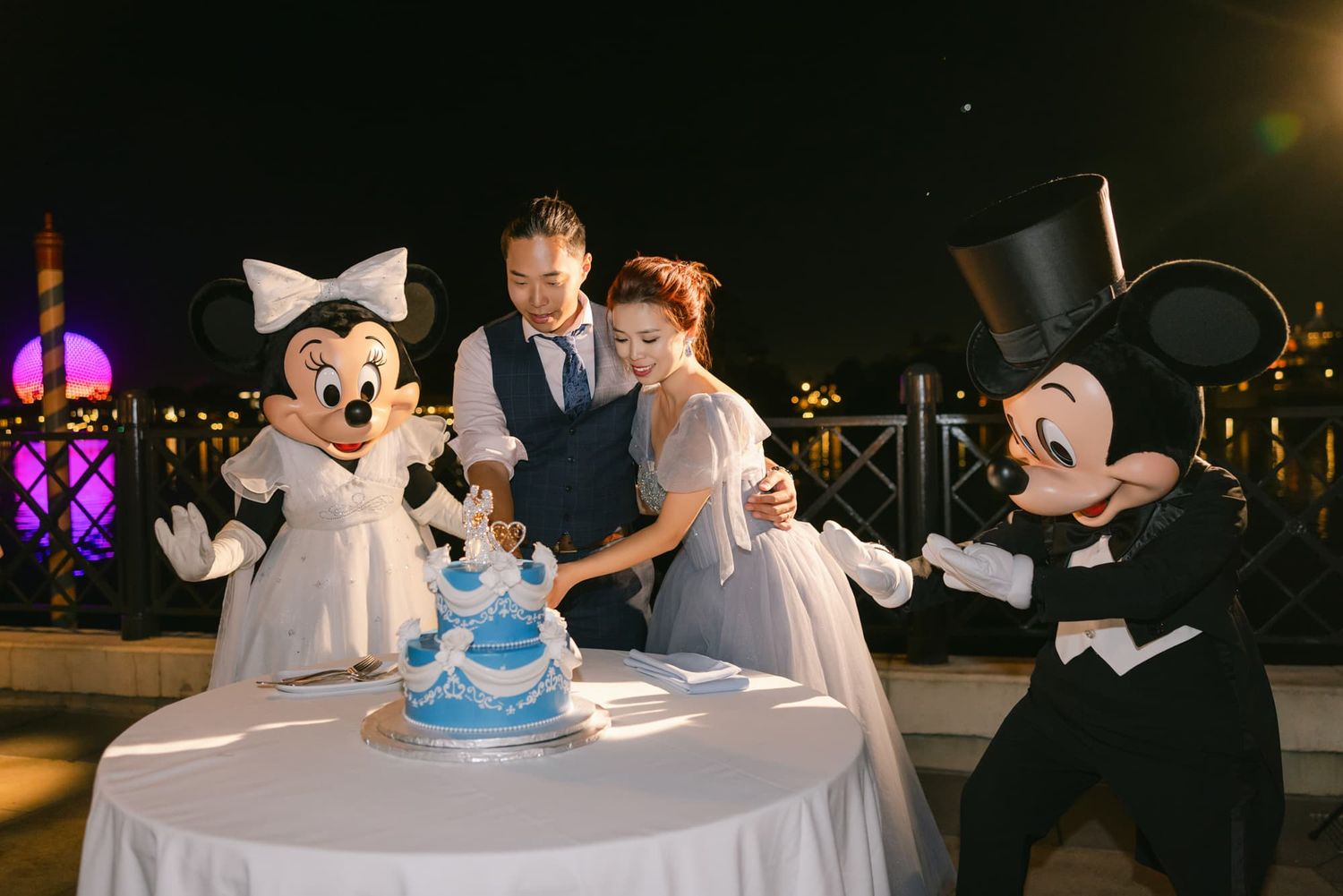Couple cuts wedding cake with Mickey and Minnie Mouse characters at a Disney-themed wedding reception at night.