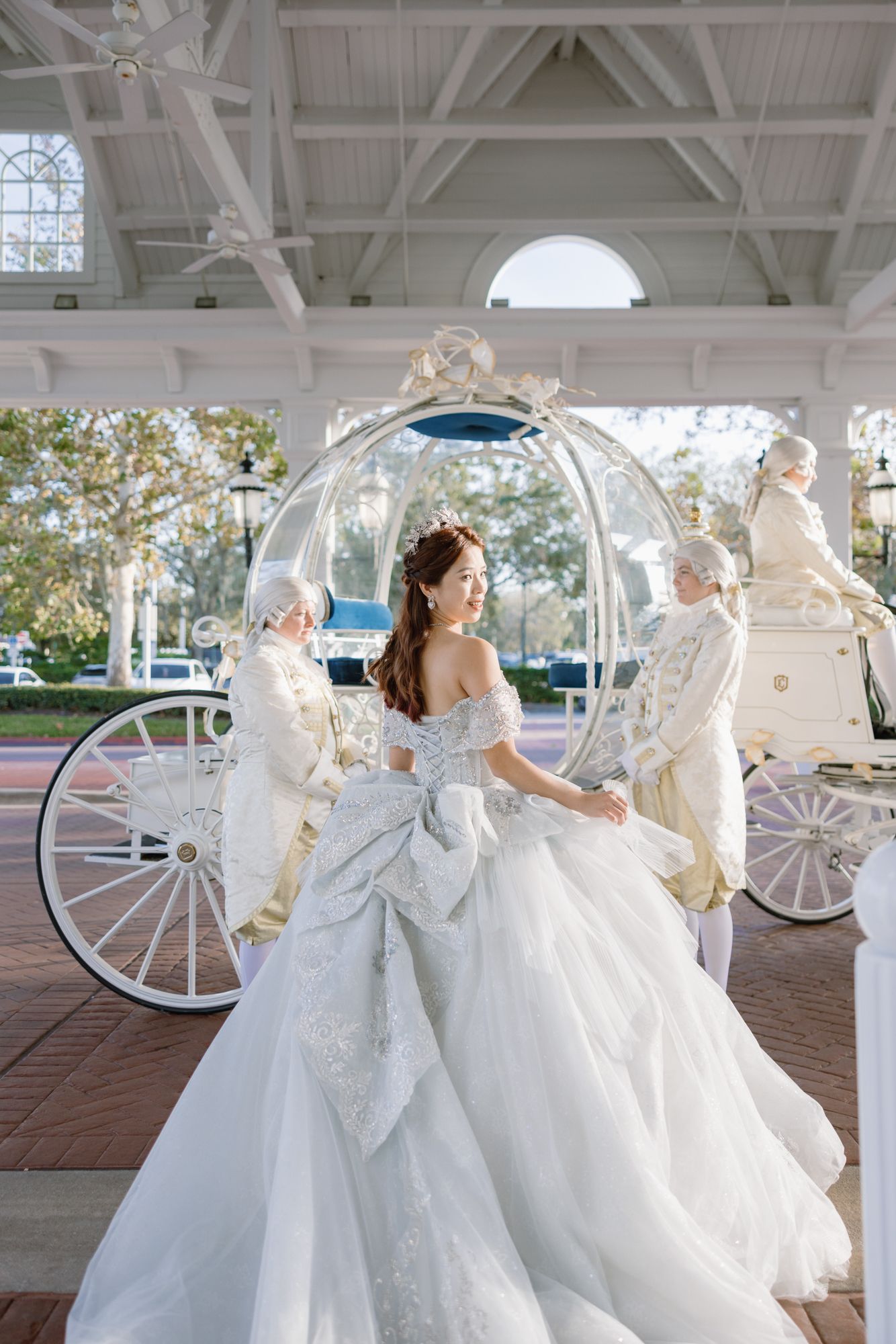 Bride in stunning off-shoulder ball gown stands before a Cinderella glass carriage at Disney World wedding.