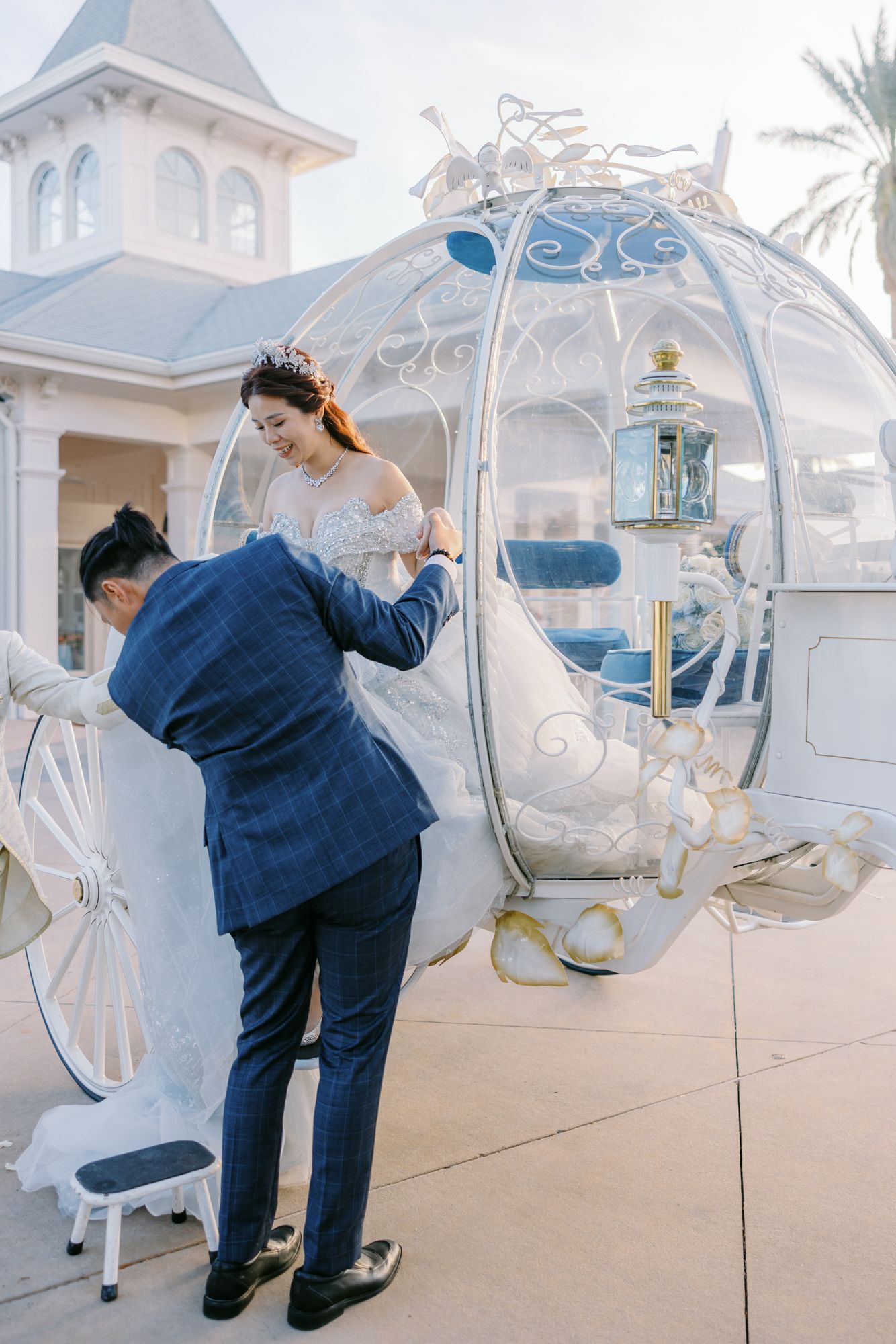 Groom in navy plaid suit helps bride into ornate Cinderella-style white carriage at Disney wedding.