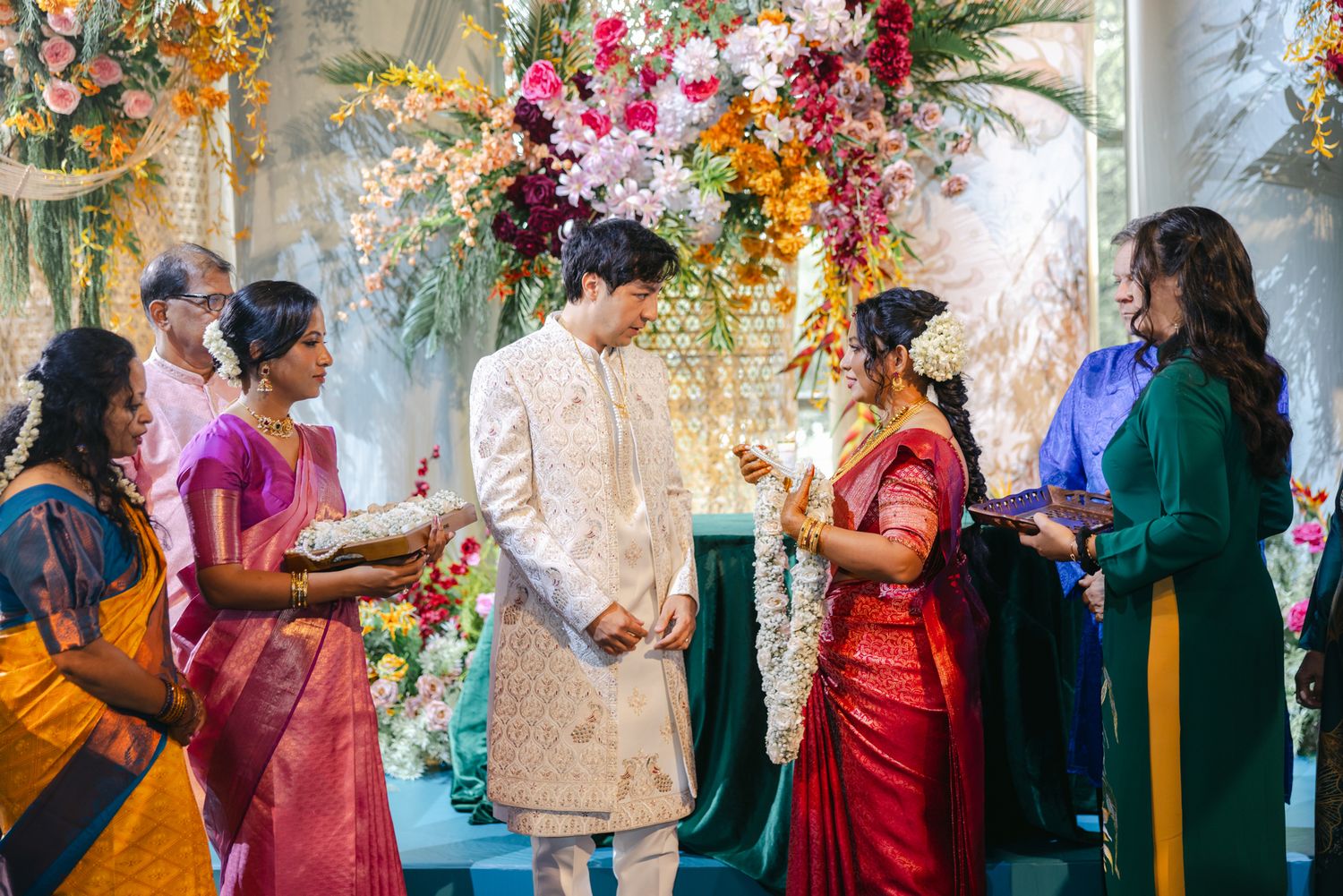 Groom in a cream sherwani and bride in a red and white saree exchange garlands during a colorful Indian wedding ceremony.