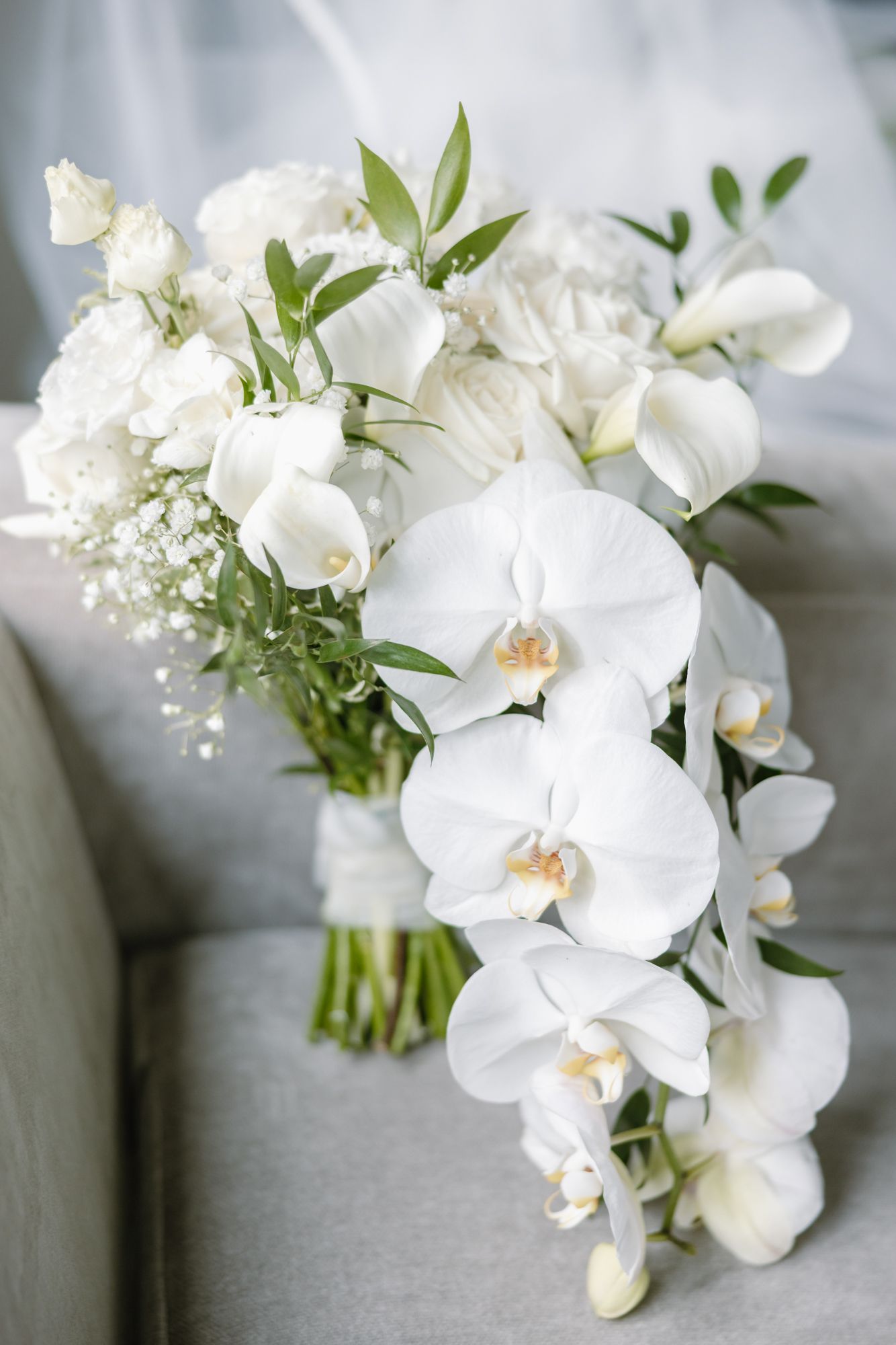 White orchid and rose cascading bridal bouquet with baby's breath and greenery resting on a gray upholstered couch.