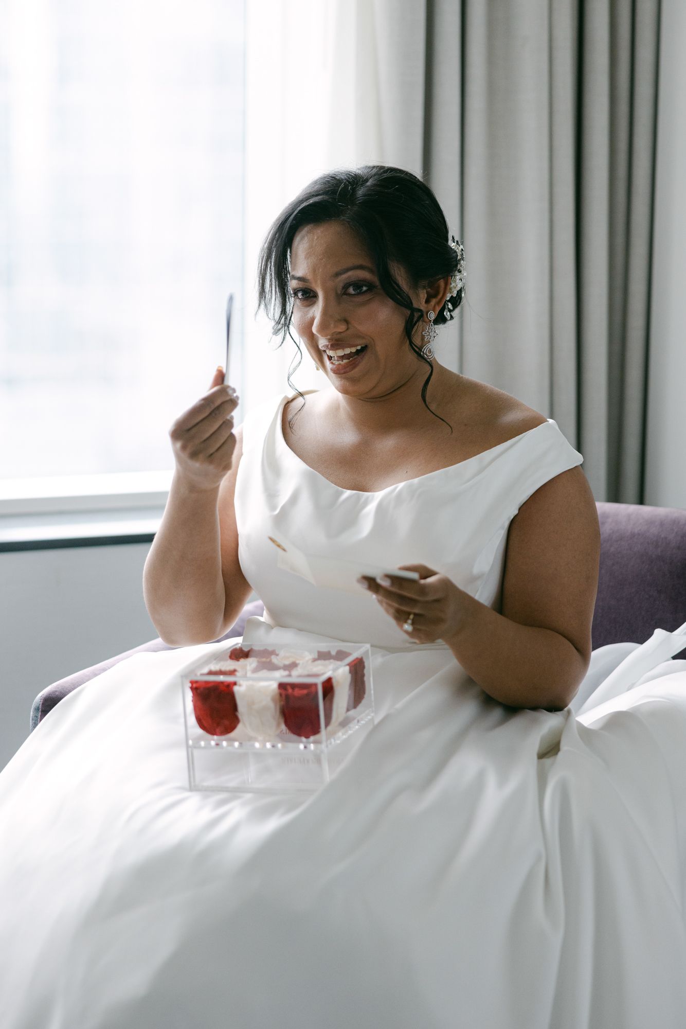 Smiling bride in an elegant white gown sits by a sunlit window, reading a card beside a clear box of red and white roses.
