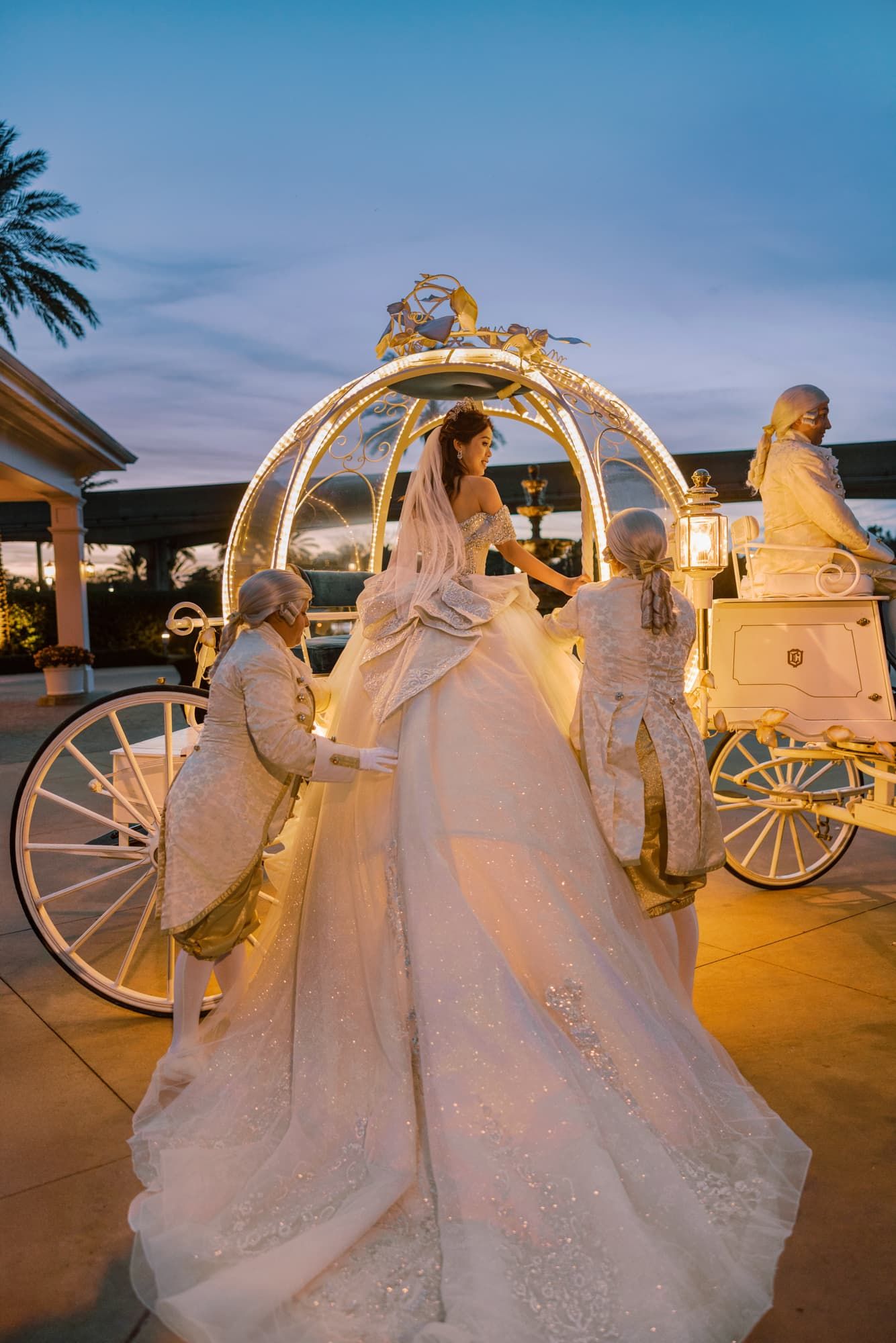 Bride in a white ballgown entering a glowing Cinderella carriage at dusk, assisted by attendants in formal attire.