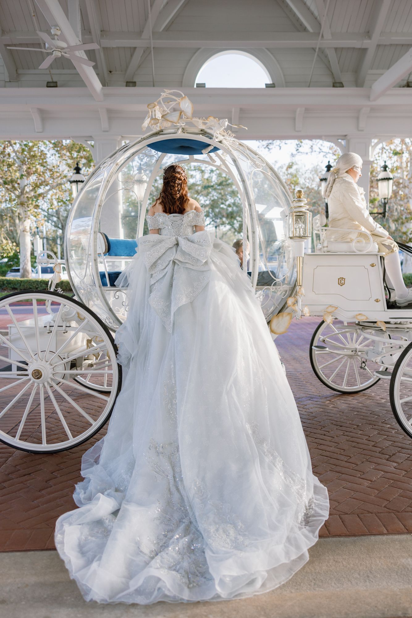 Bride in white lace ball gown standing before Cinderella glass carriage from behind.