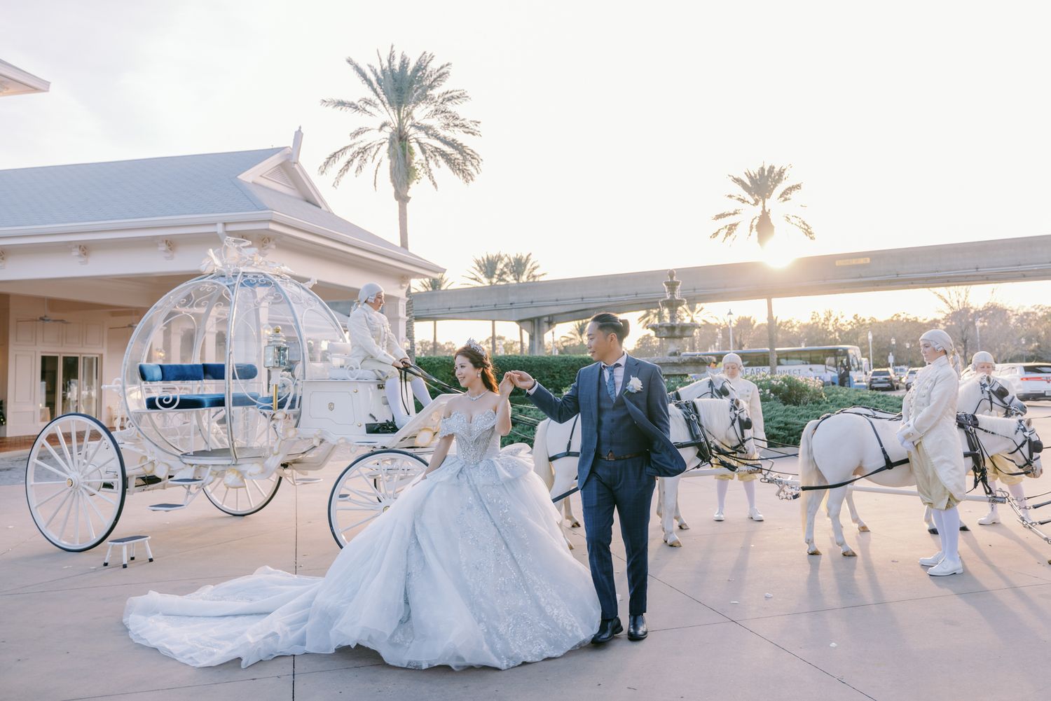 Bride and groom posing with white Cinderella carriage and horses at golden sunset.