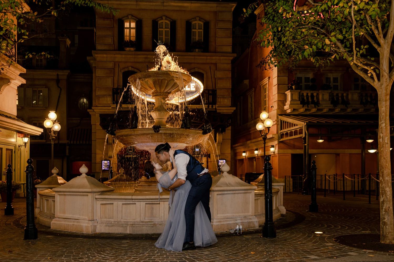 Couple sharing romantic dip kiss in front of illuminated fountain at night in elegant outdoor plaza.