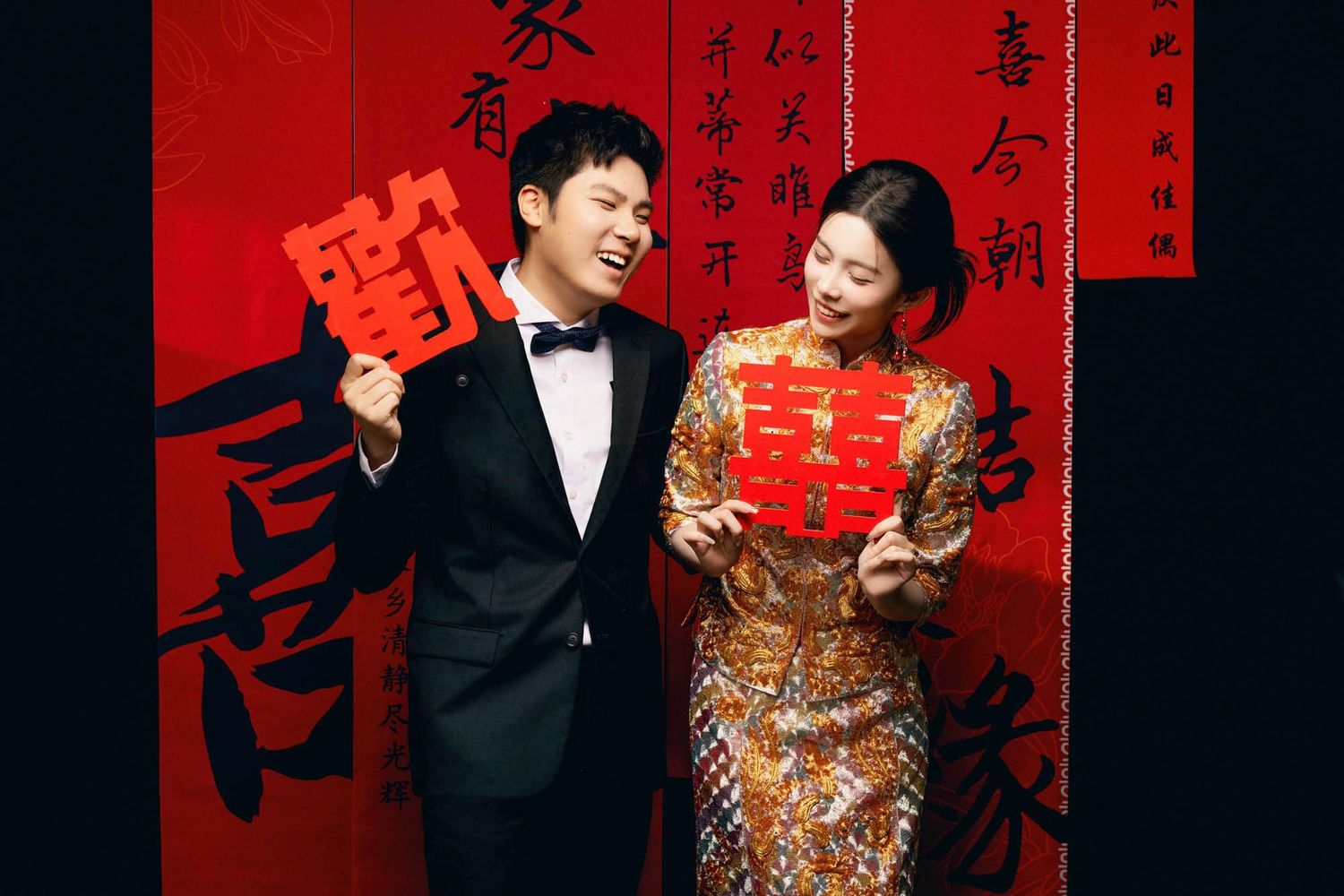 Chinese couple in traditional wedding attire holding red double happiness symbol against red calligraphy backdrop.