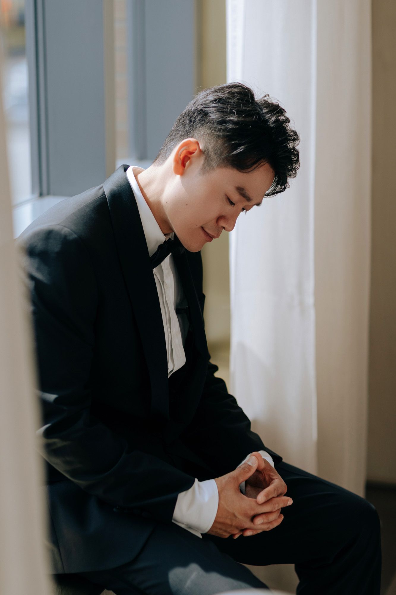 Contemplative groom in black tuxedo sitting by sunlit window before wedding ceremony.