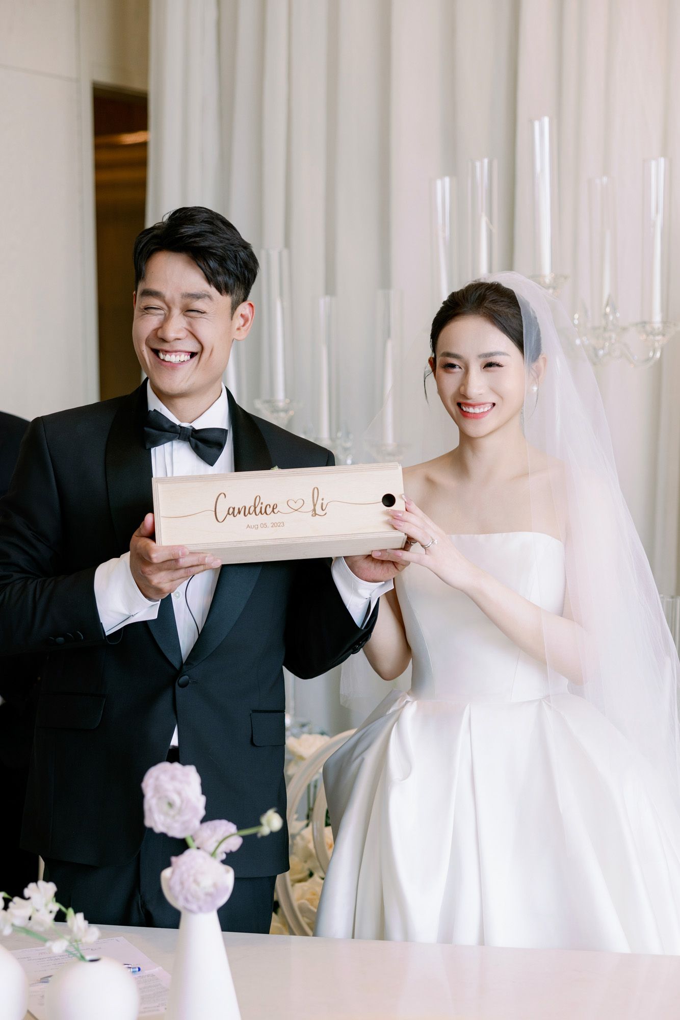 Smiling bride and groom hold personalized wooden keepsake box at elegant wedding reception table.
