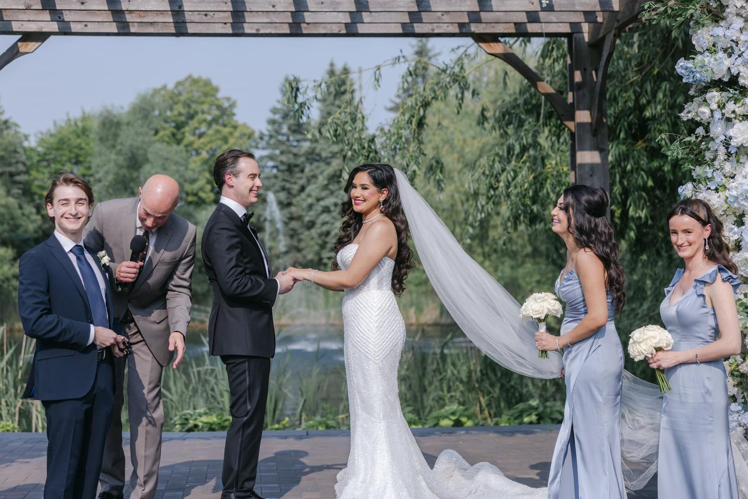 Wedding party stands together at outdoor ceremony beside scenic pond and pergola backdrop.