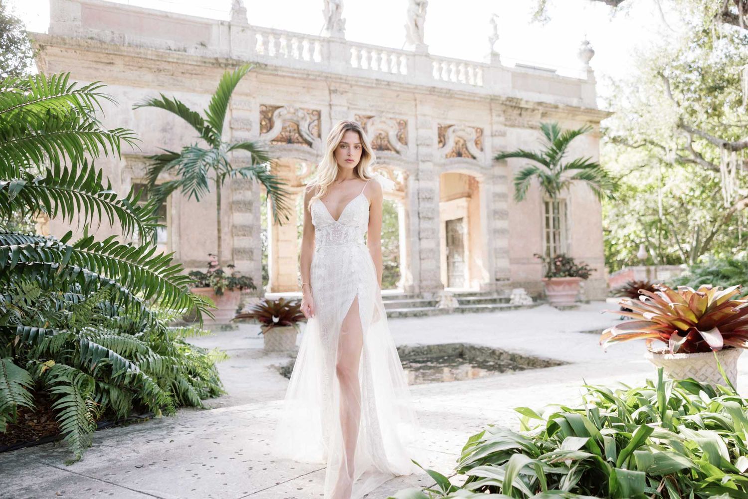 A bride in a lace wedding dress with a thigh-high slit stands in a lush garden courtyard.