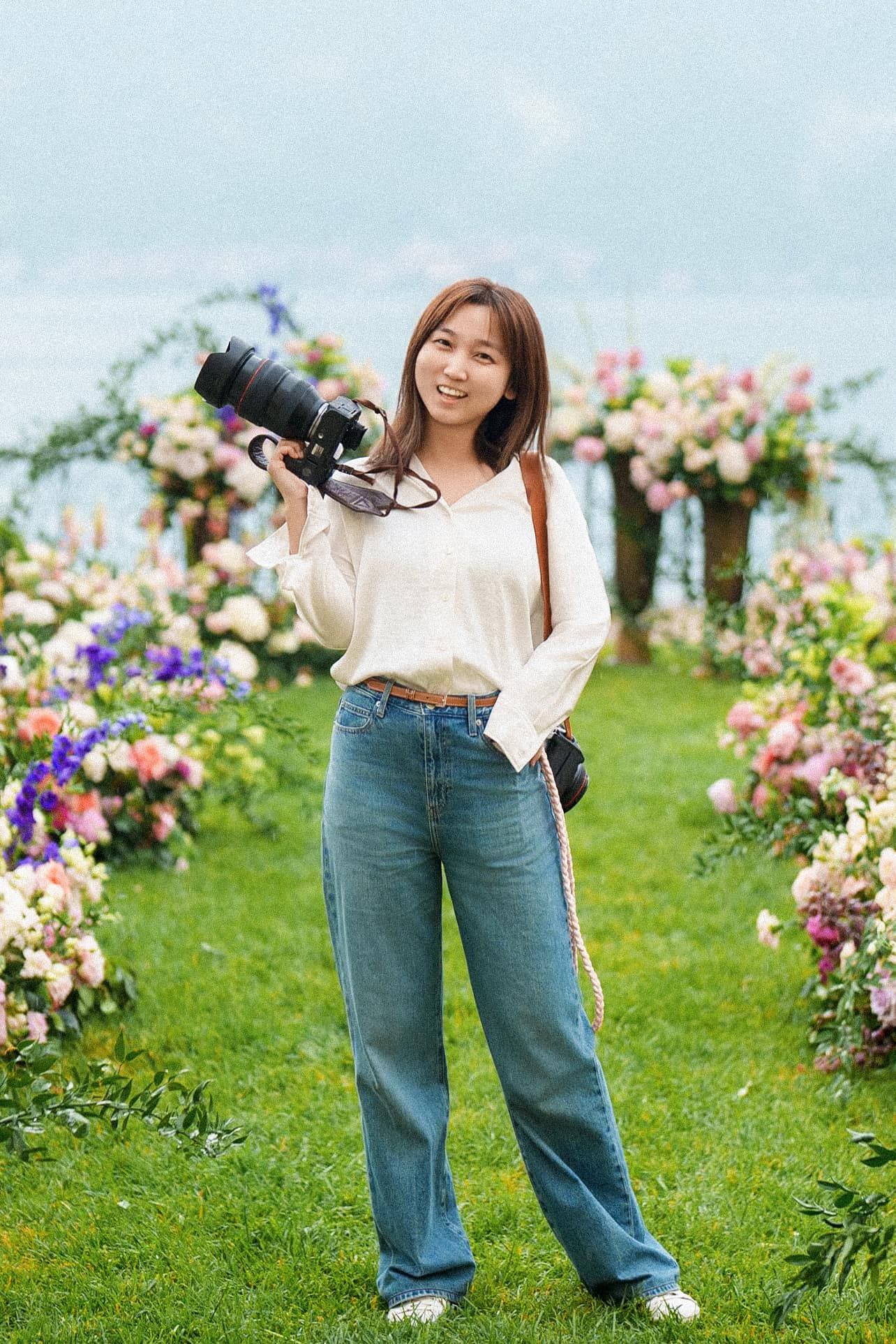 Smiling female photographer holding camera in a lush floral garden setting.
