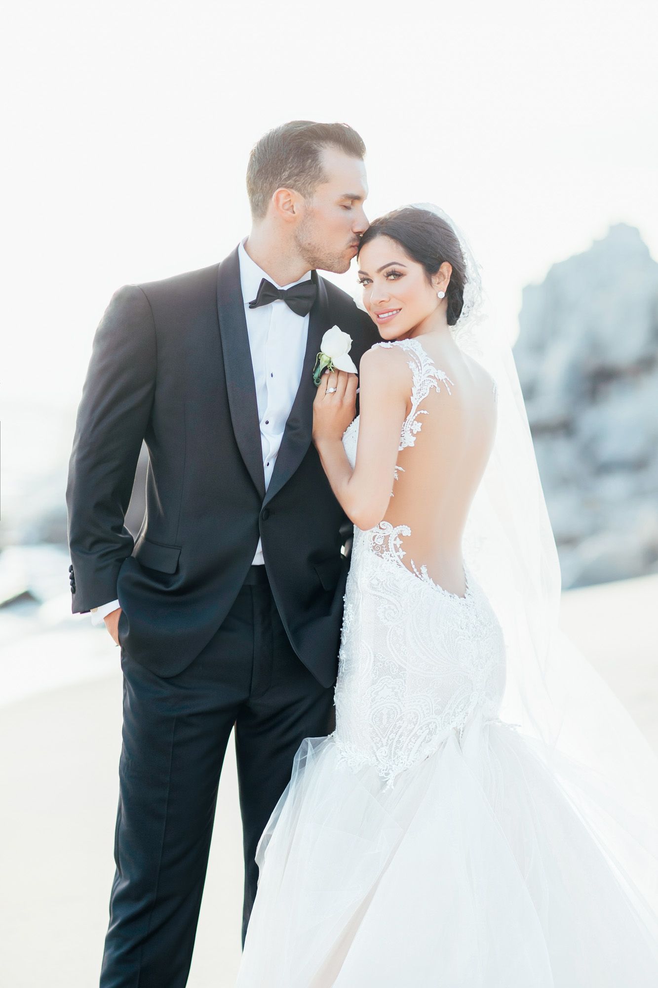 Groom kisses bride on the forehead as she looks back, showcasing her lace backless mermaid wedding gown.