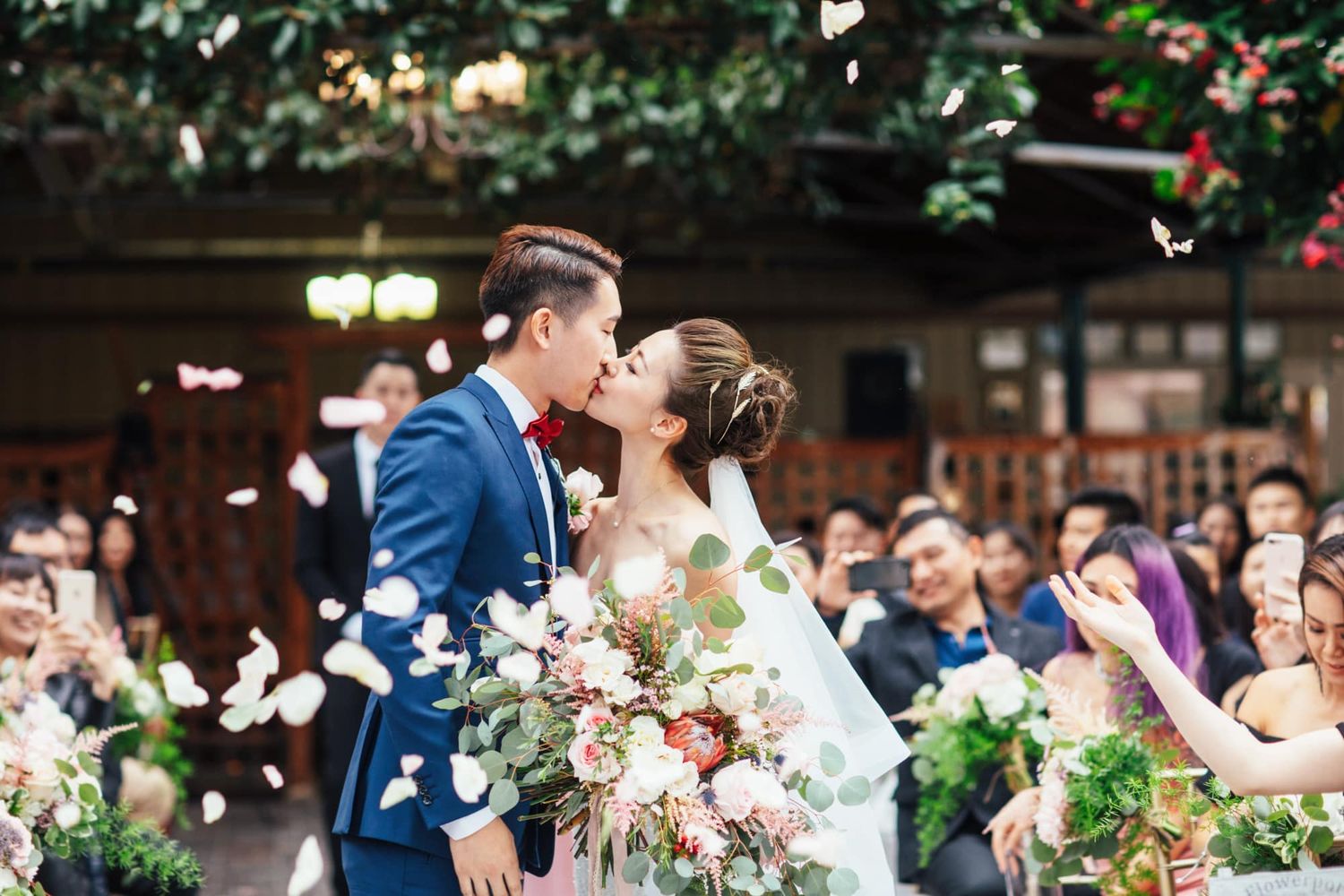 Bride and groom share a kiss at outdoor wedding ceremony as guests toss white flower petals in celebration.