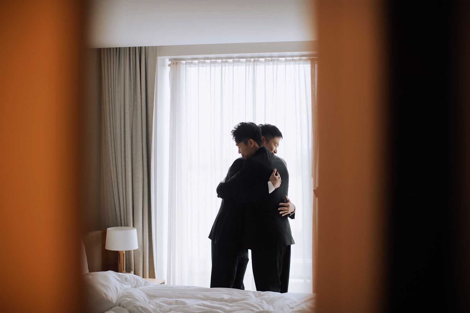 Two men in black suits share an emotional embrace by a sunlit hotel room window.
