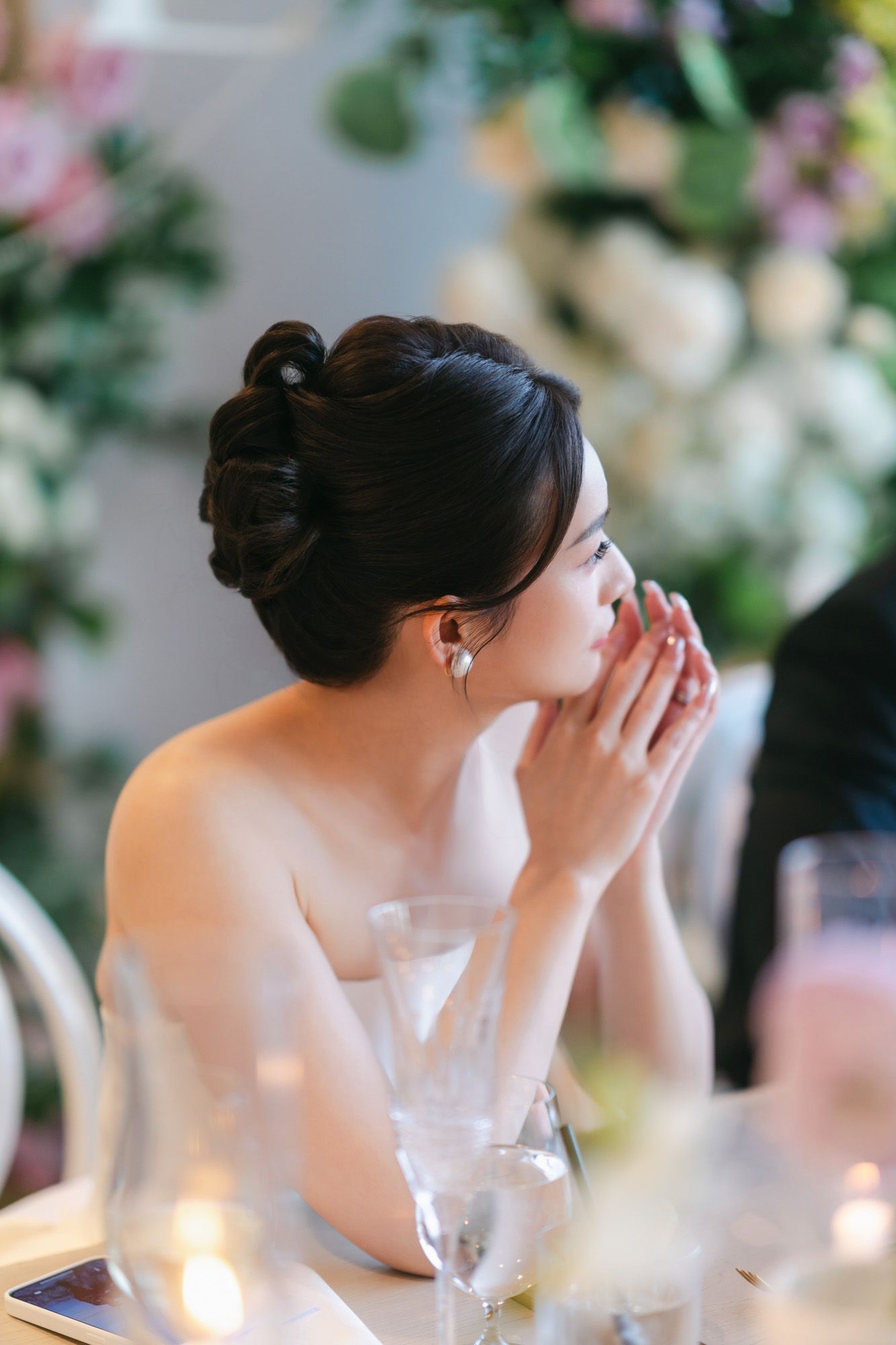 Elegant bride with updo hairstyle sitting at wedding table surrounded by floral arrangements.