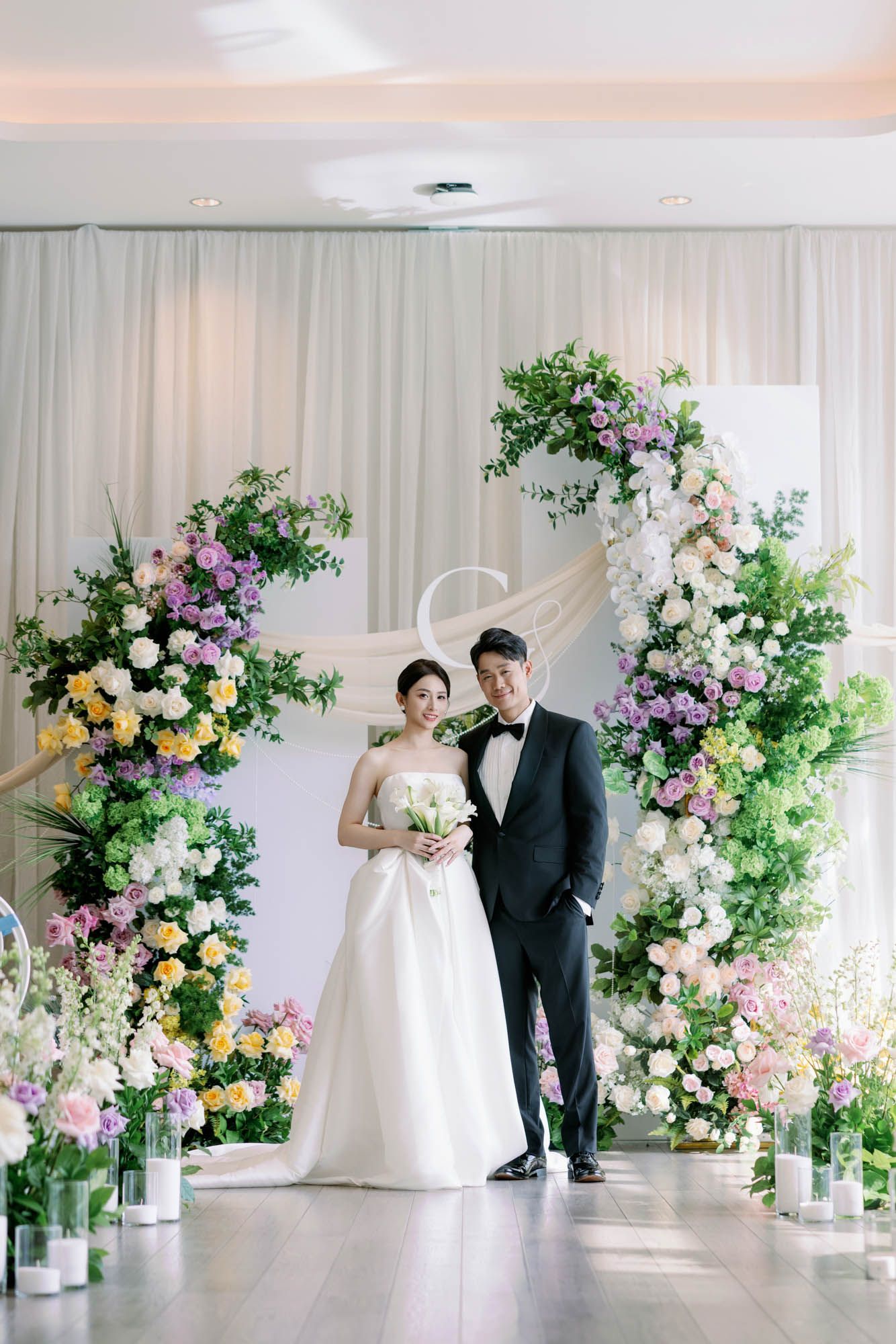 Bride and groom pose before stunning floral arch with colorful blooms at indoor wedding ceremony.