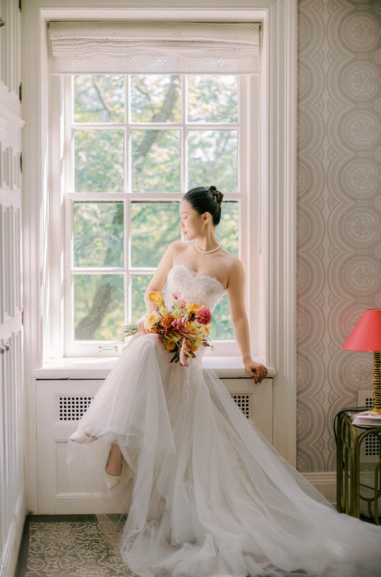 Bride in strapless tulle gown sitting by a window holding a vibrant floral bouquet.