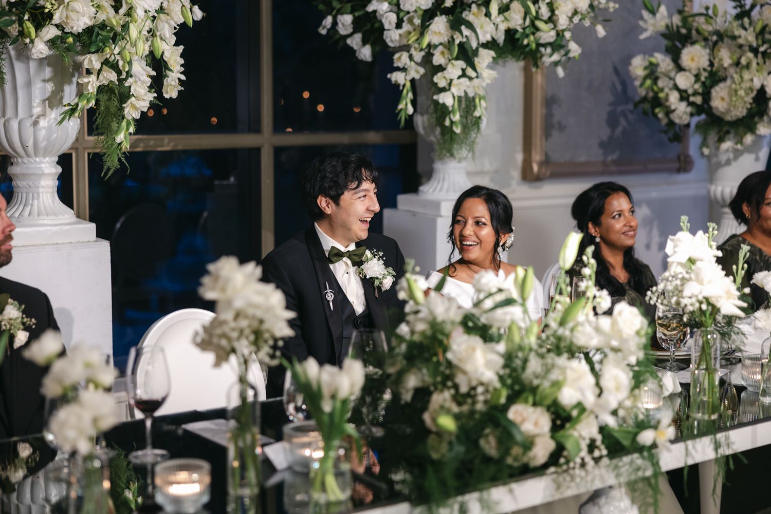 Bride and groom smile at the head table surrounded by white floral centerpieces and wedding guests during the reception.