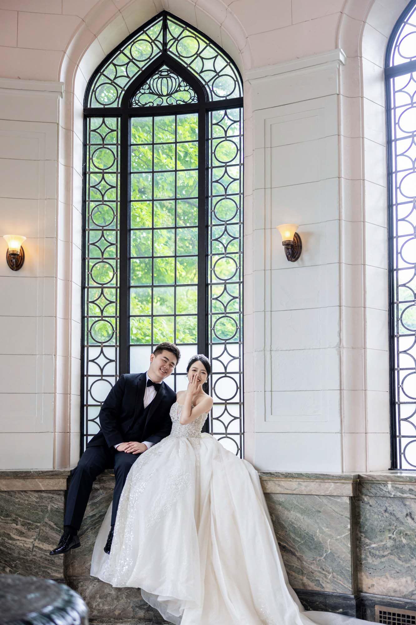 Bride and groom sitting together by an ornate arched stained glass window in an elegant white chapel.
