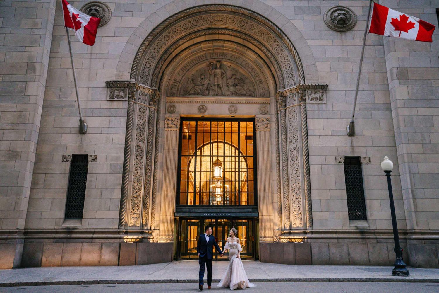 Bride and groom stand before a grand stone building entrance with Canadian flags at a Ottawa wedding.