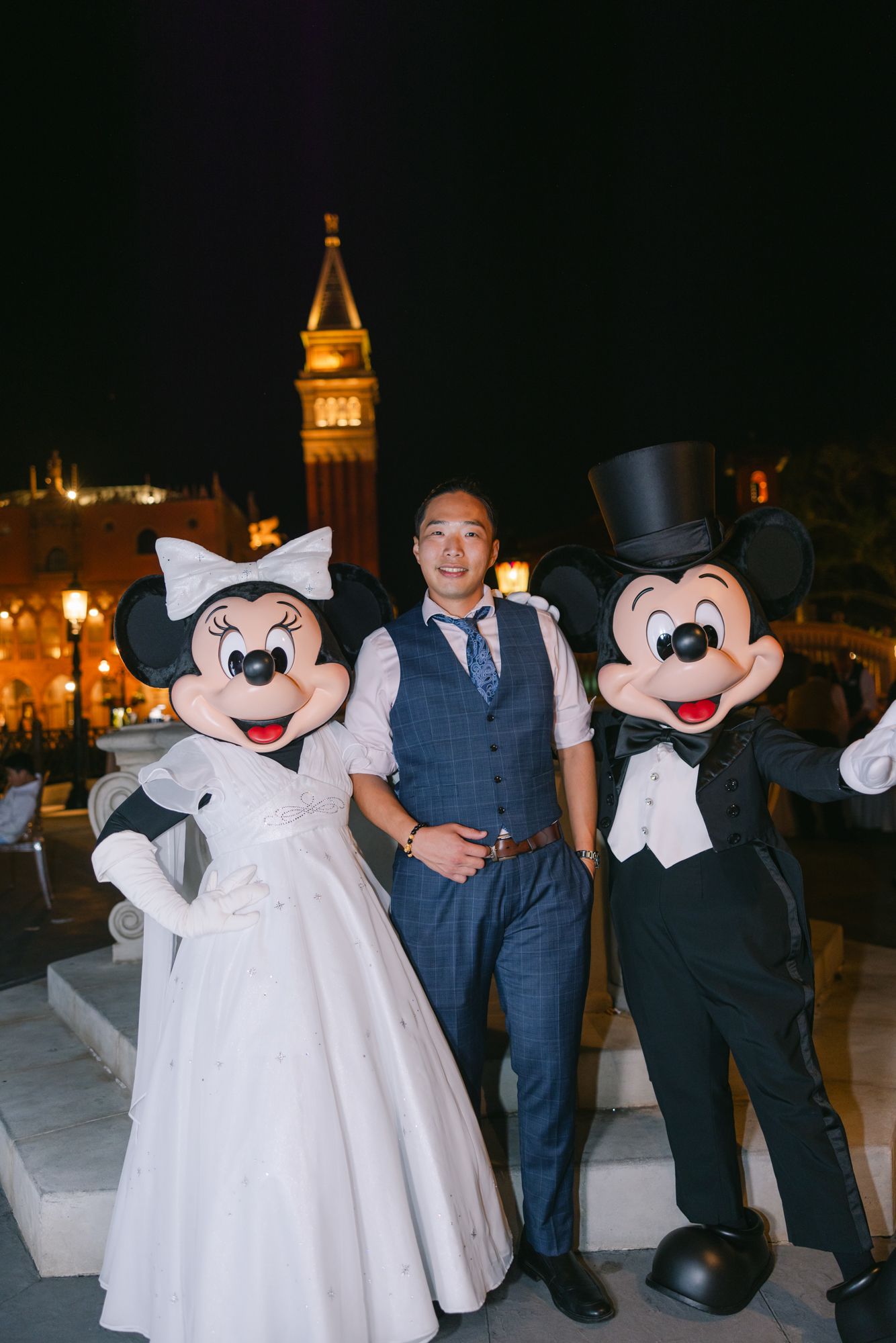 Groom poses with Minnie and Mickey Mouse in wedding attire at Disney World at night.