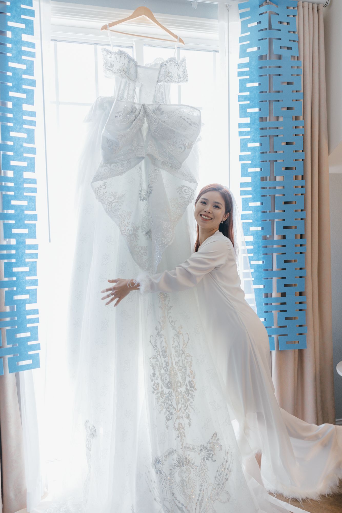 Smiling bride in white robe embracing her elegant lace wedding gown hanging by a bright window.
