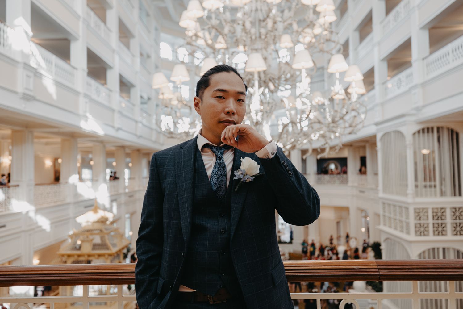 Handsome groom in navy suit with floral boutonniere poses thoughtfully in ornate chandelier-adorned grand atrium.