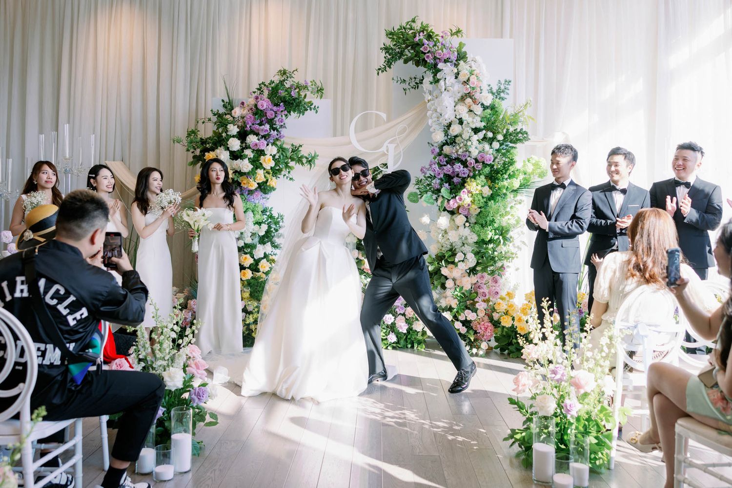 Groom dramatically dips bride before floral arch as wedding guests cheer and photograph the moment.