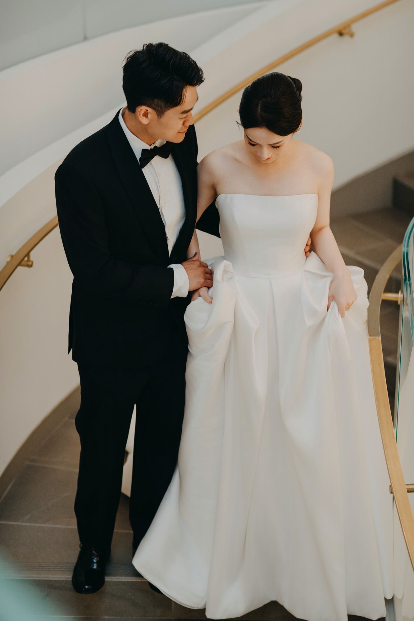 Groom in black tuxedo assists bride in strapless white ball gown on a spiral staircase.