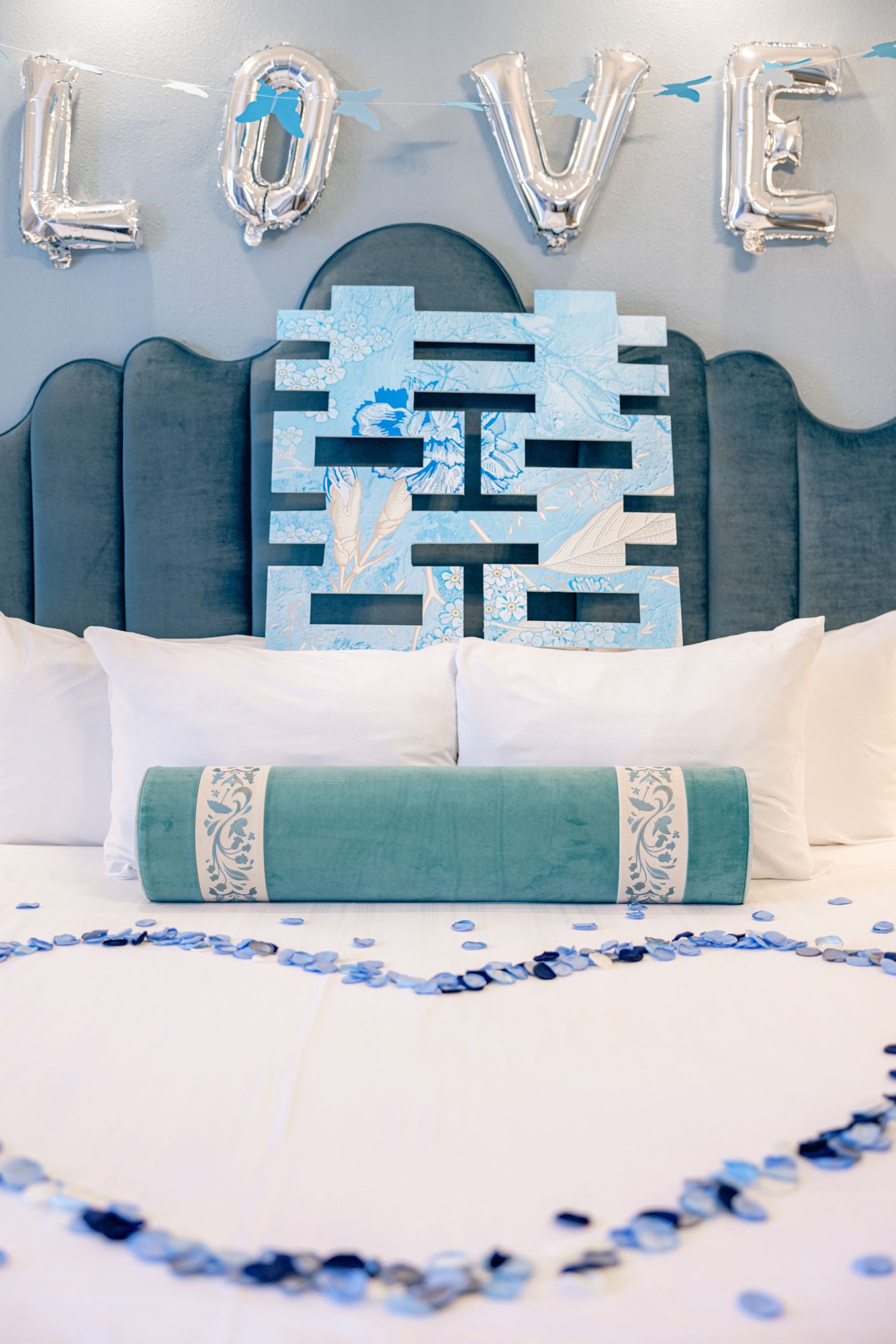 Blue wedding bedroom decorated with LOVE balloons, double happiness symbol, teal bolster pillow, and heart-shaped confetti petals.