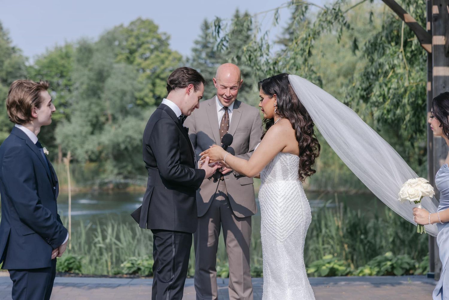 Groom placing wedding ring on bride's finger during outdoor ceremony by a serene lake.