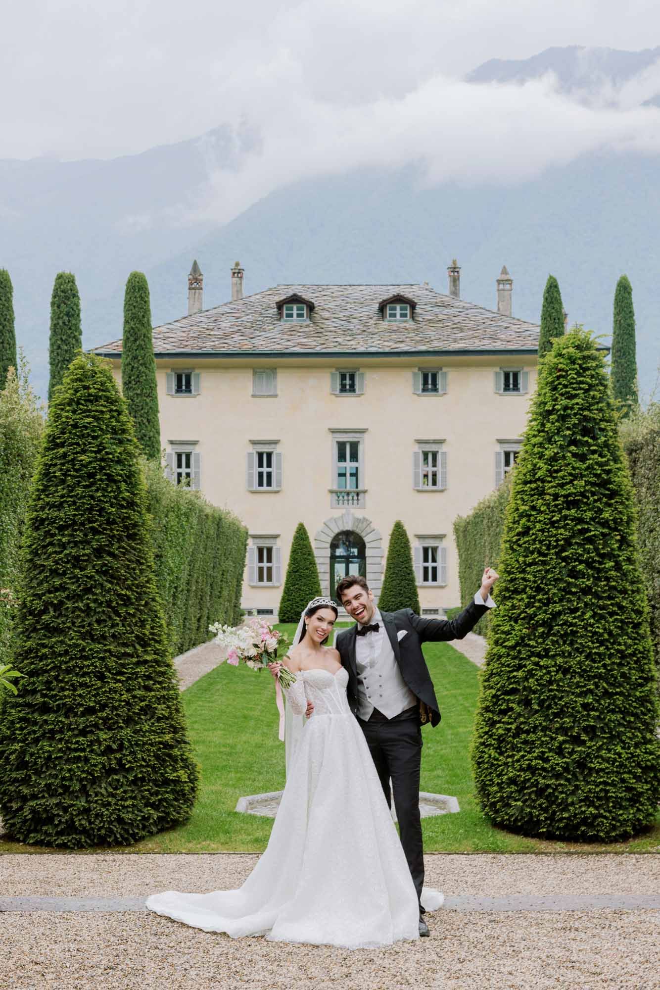 Bride and groom celebrate joyfully in formal gardens with cypress trees and an elegant Italian villa backdrop.