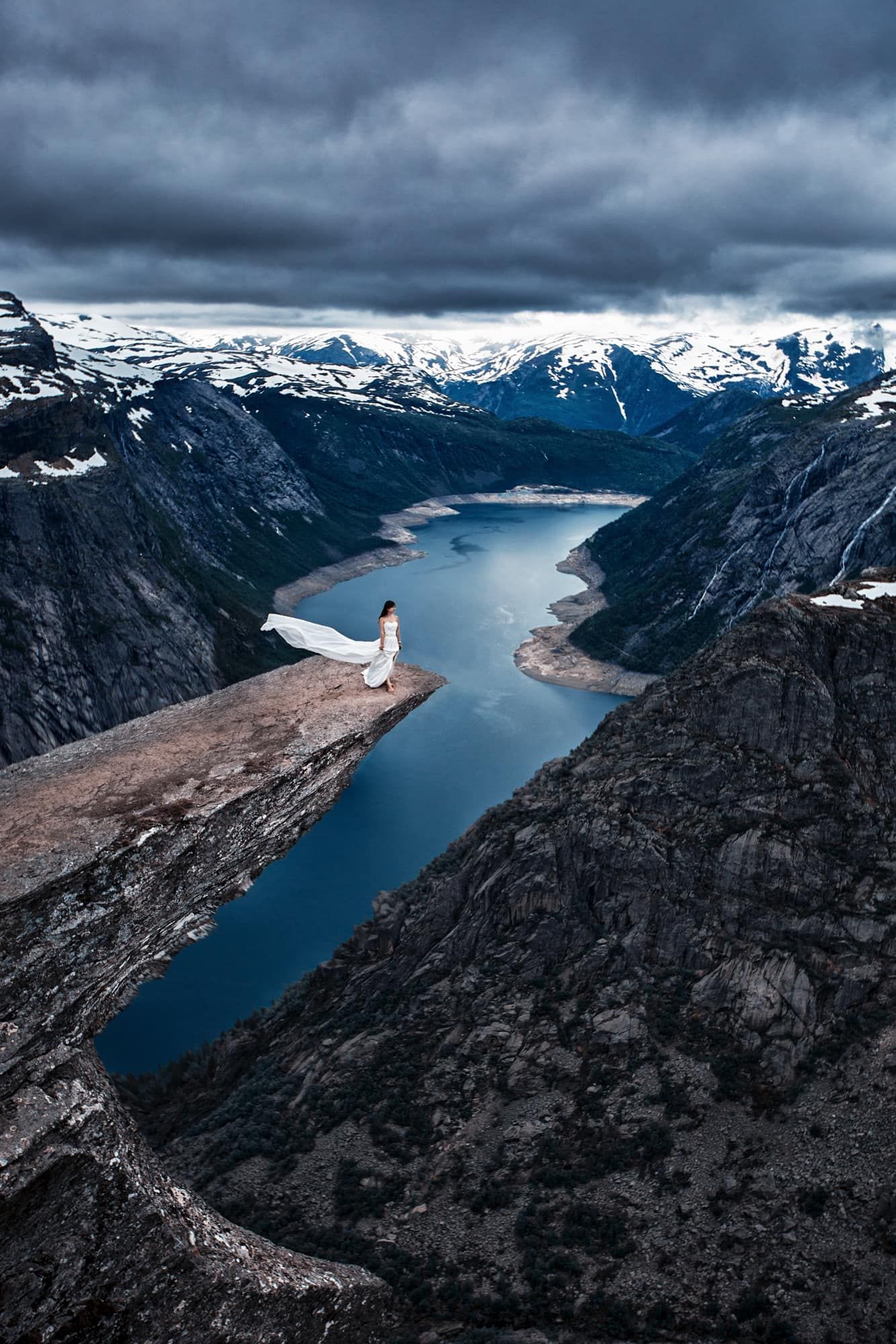 Bride in white gown standing on Trolltunga cliff edge overlooking stunning Norwegian fjord landscape.