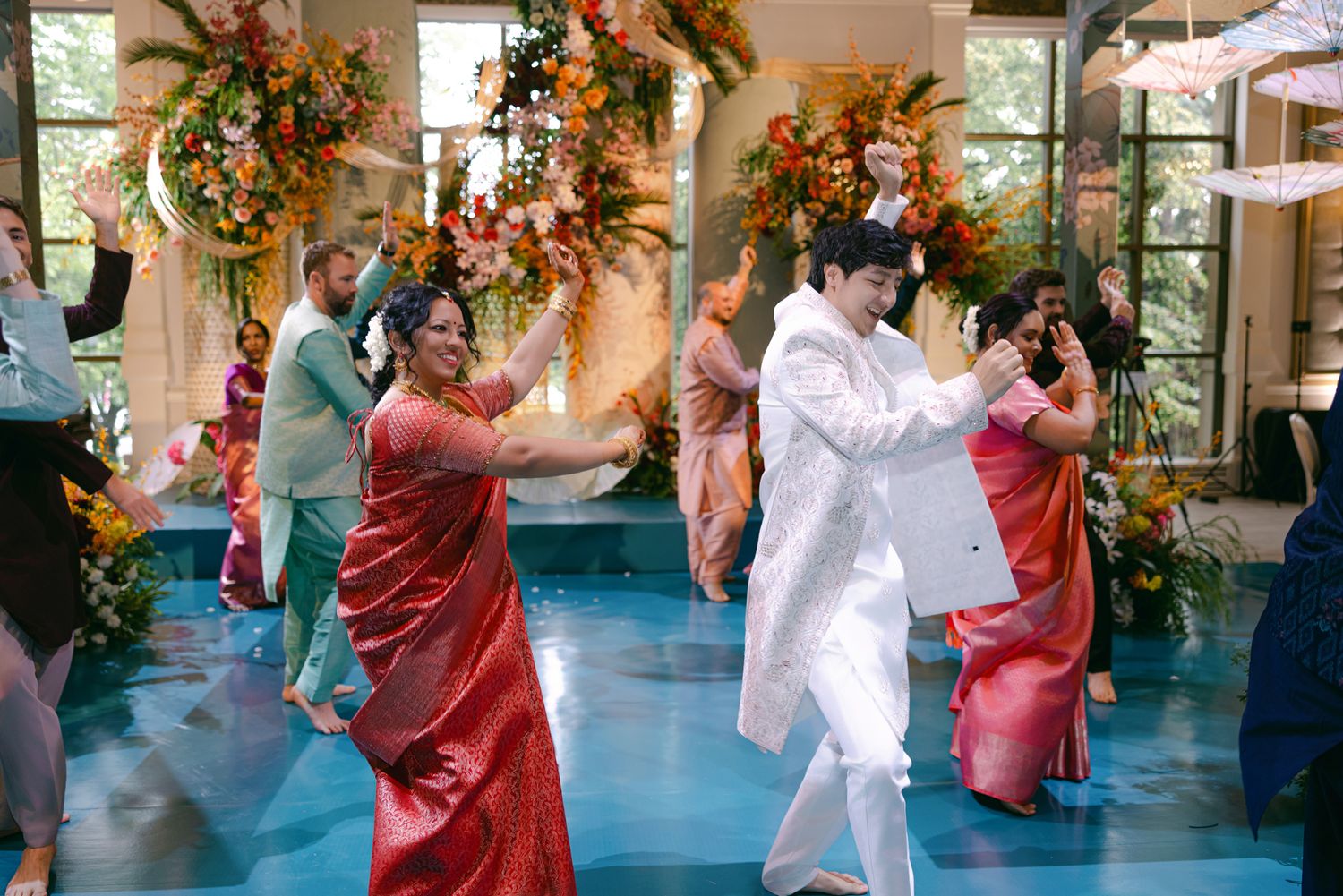 Wedding guests in colorful traditional outfits dance joyfully with the groom in a floral decorated venue with a blue floor.