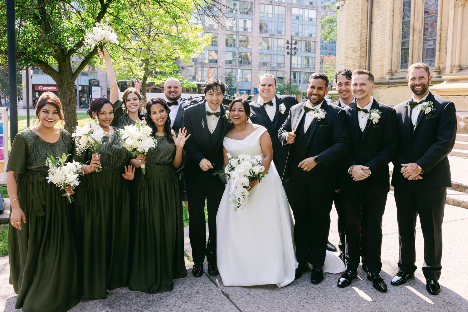 Bride in a white gown surrounded by bridesmaids in green dresses and groomsmen in dark suits posing outdoors on a city sidewalk.