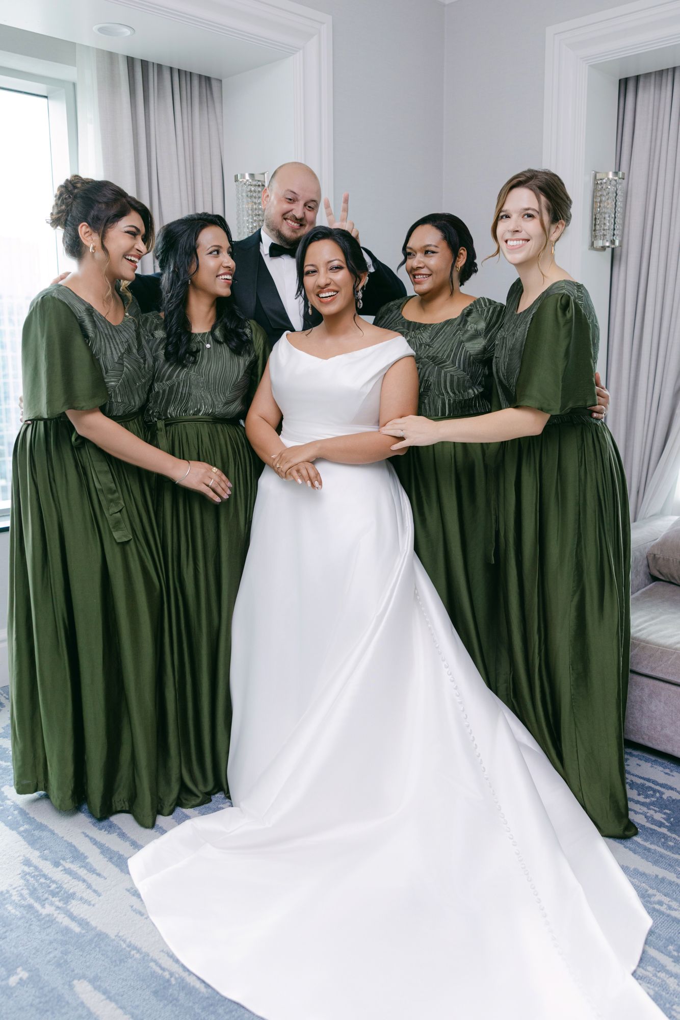 Bride in a white satin gown smiles with four bridesmaids in olive green dresses and a groomsman in a tuxedo.