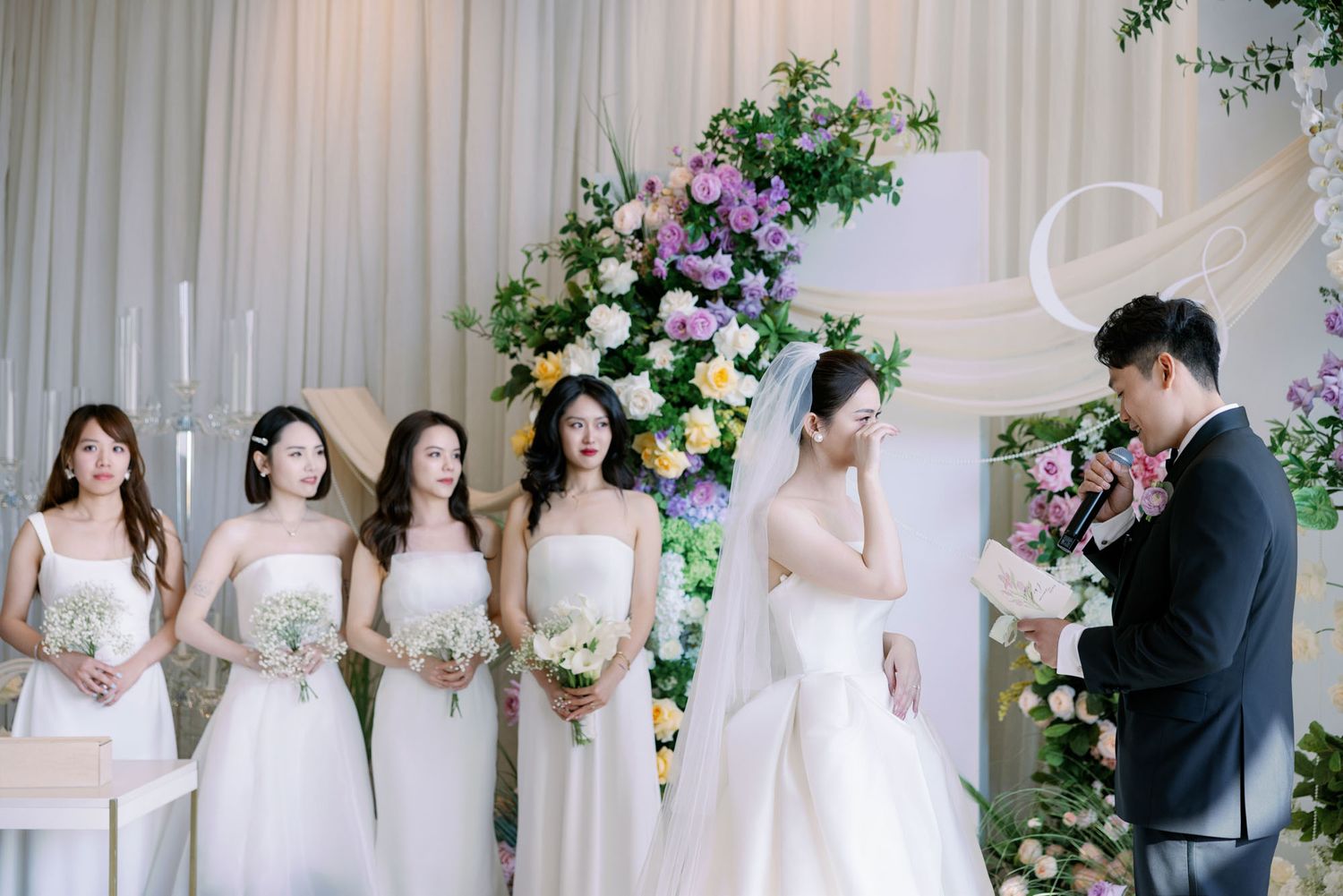 Bridesmaids in white dresses hold bouquets beside bride and groom during wedding ceremony.