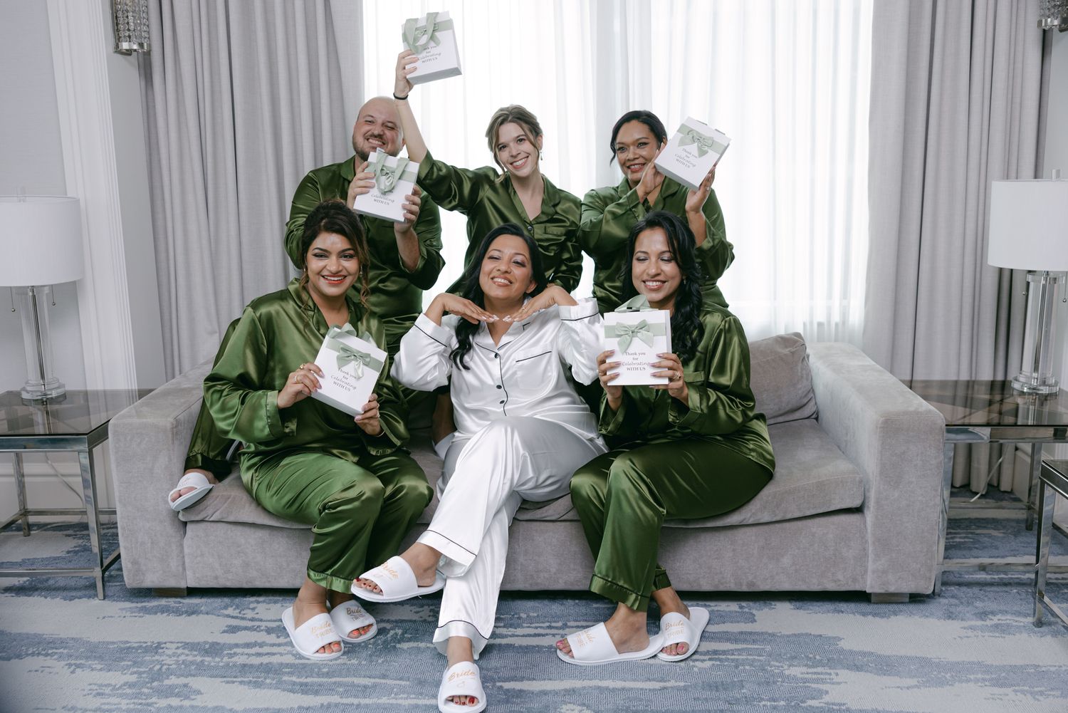 Six bridesmaids in matching green satin pajamas and white slippers sit on a gray sofa, smiling and holding up gift boxes.