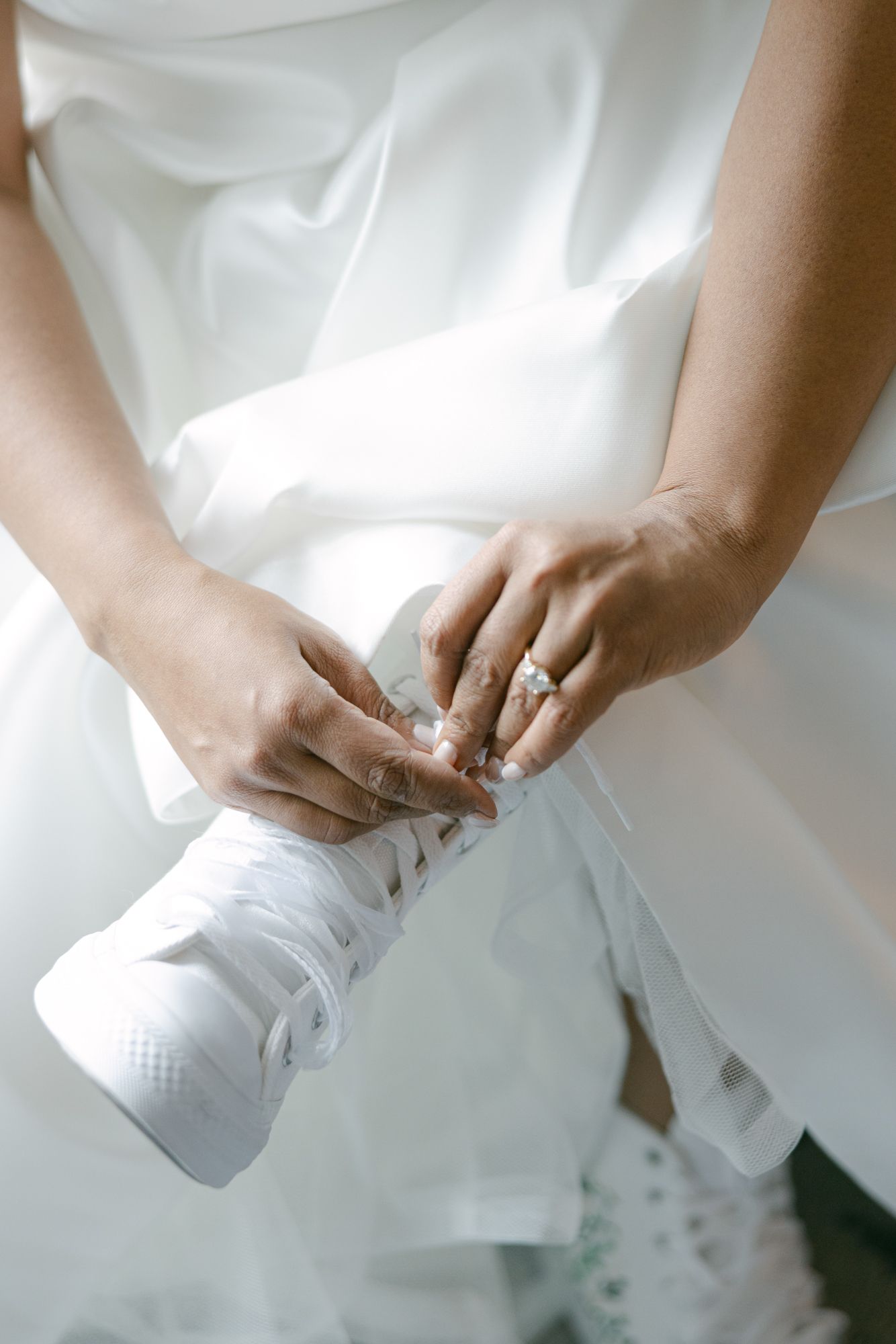 Bride in a white wedding gown laces up a white sneaker while wearing a sparkling ring on her finger.
