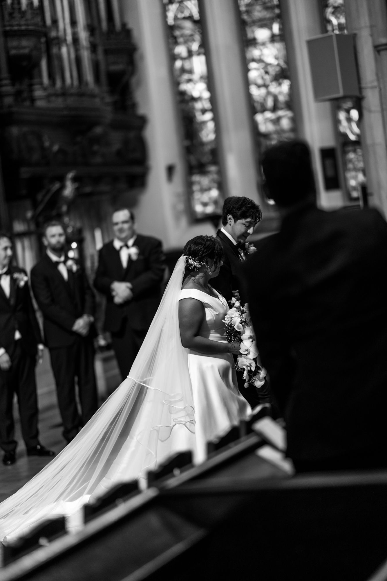 Bride in a white gown and long veil holds a bouquet at the altar of a stone church while groomsmen in black suits look on.