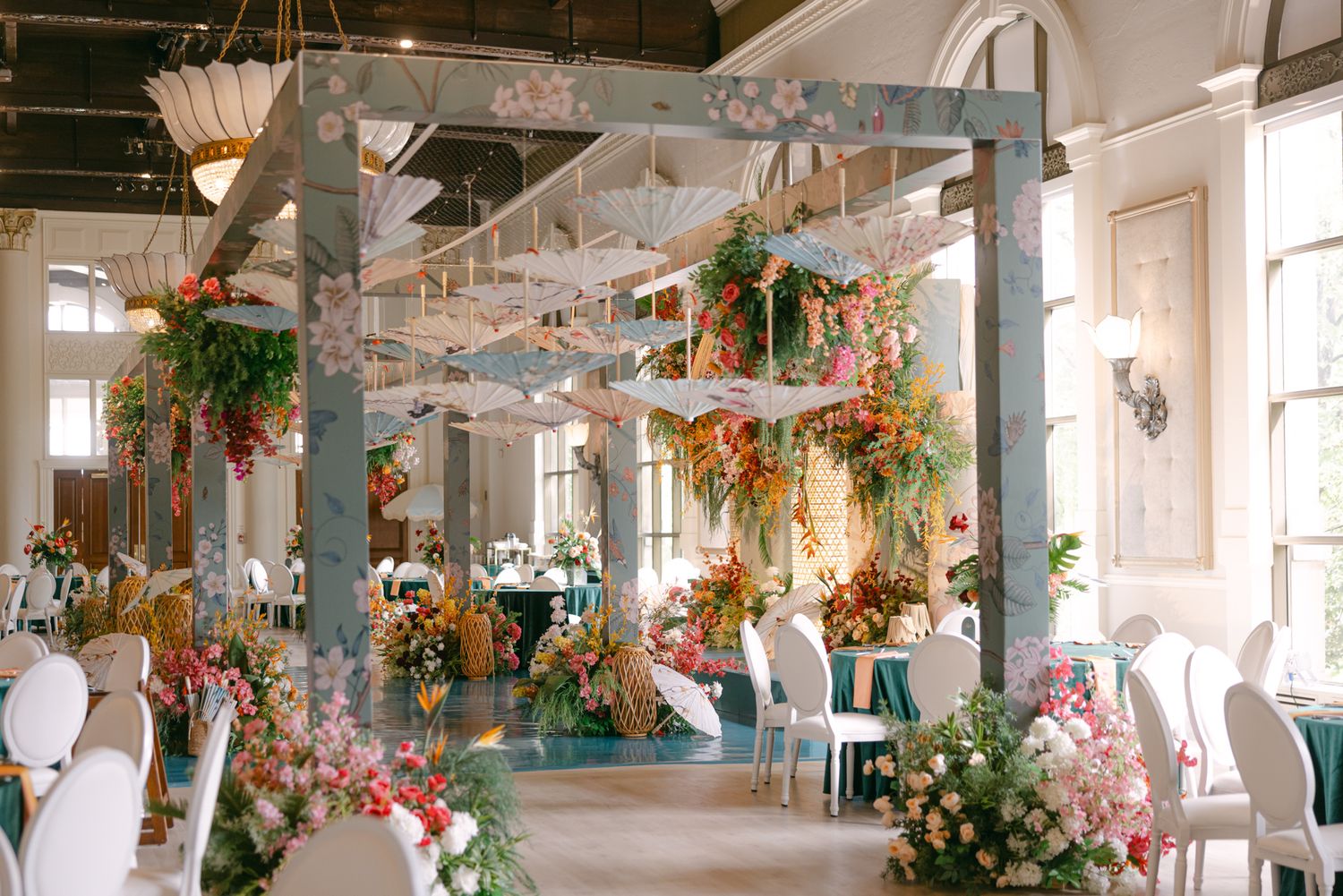 Wedding reception hall decorated with a floral archway, hanging umbrellas, and colorful bouquets above tables and white chairs.