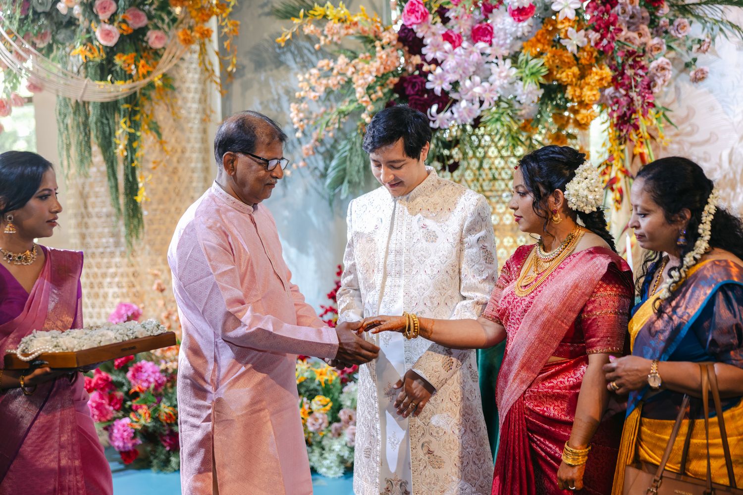 Family members in pink, white and silk saris exchange a ceremonial gesture at an Indian wedding with colorful floral decor.