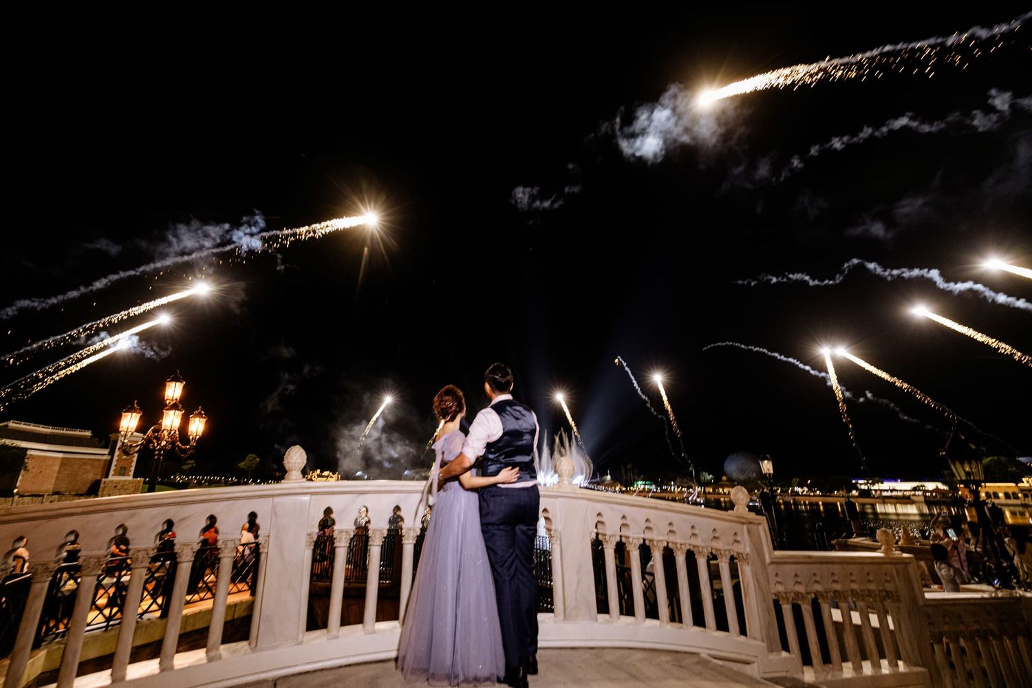 Couple embraces on elegant balcony with stunning fireworks display lighting the night sky at a romantic wedding celebration.