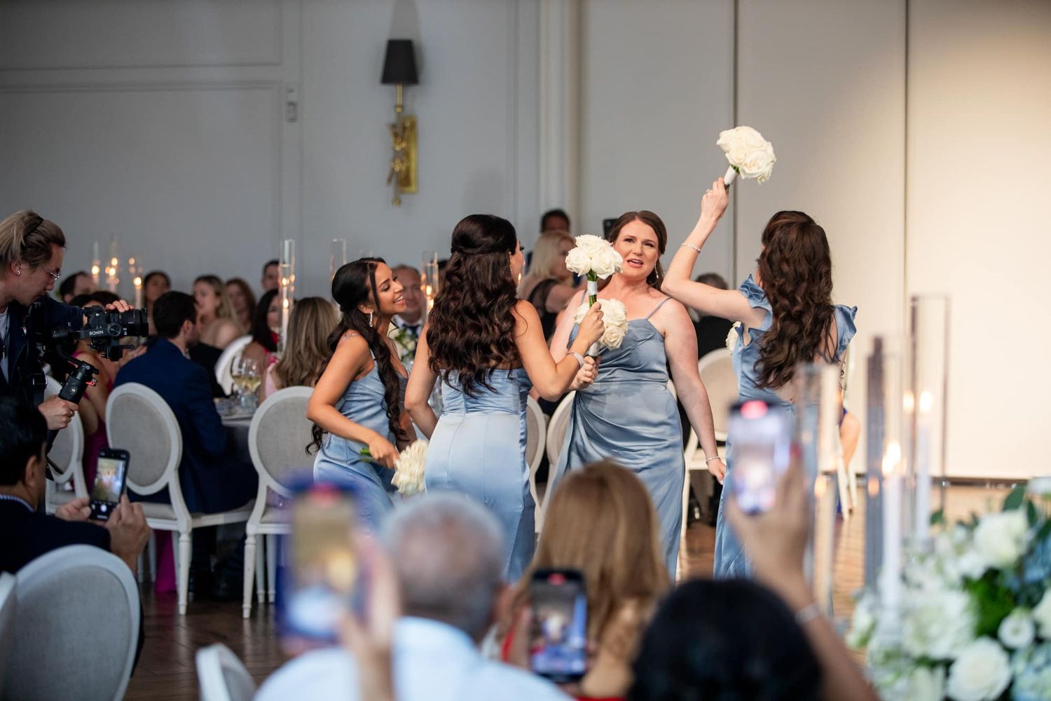 Bridesmaids in blue gowns walking down the aisle carrying white bouquets at a wedding reception.